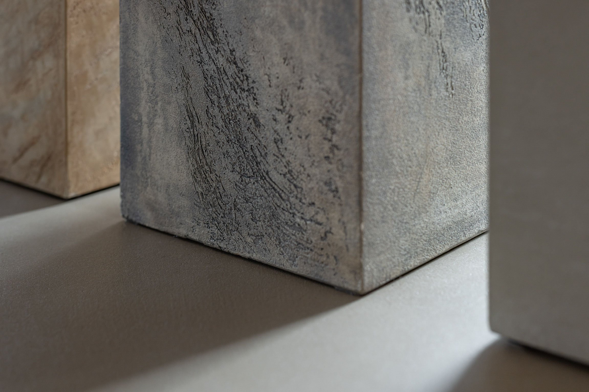 Nahaufnahme von drei kubischen Marmorblöcken in neutralen Tönen, die nebeneinander auf einer flachen Oberfläche stehen, mit weicher Beleuchtung und Schatten.