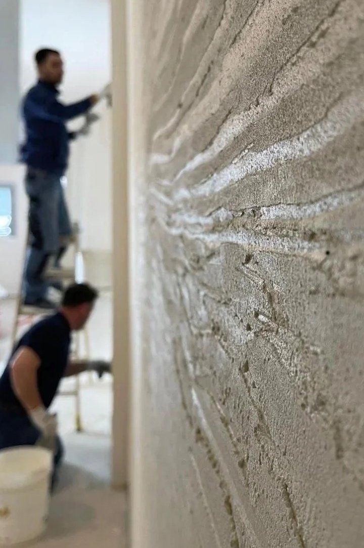 Nahaufnahme einer Wand mit Putz, im Hintergrund zwei Männer bei der Arbeit.