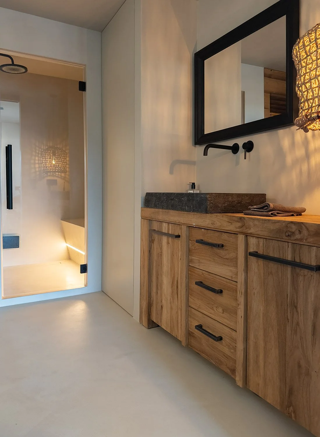 Ein Badezimmer mit Holzschrank, grauem Waschbecken, Wandspiegel, schwarzer Wasserhahn und einer geschlossenen, weißen Tür; im linken Hintergrund eine Duschkabine mit Glastür.