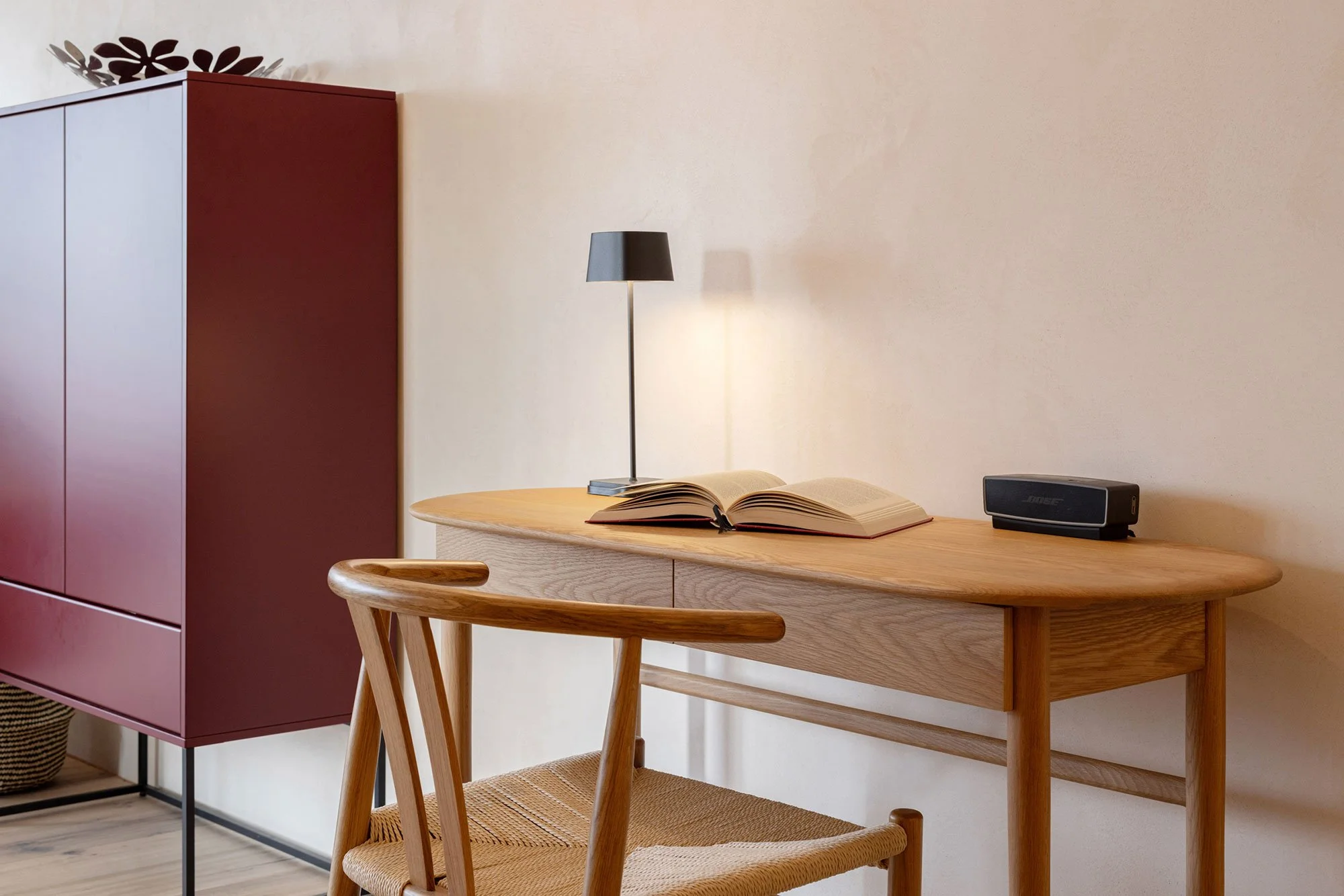 Ein Holzschreibtisch mit einem aufgeschlagenen Buch, einer Tischlampe, einem Bose-Lautsprecher und einem Stuhl steht vor einer weißen Wand. Im Hintergrund ist eine rote Schrankwand sichtbar.