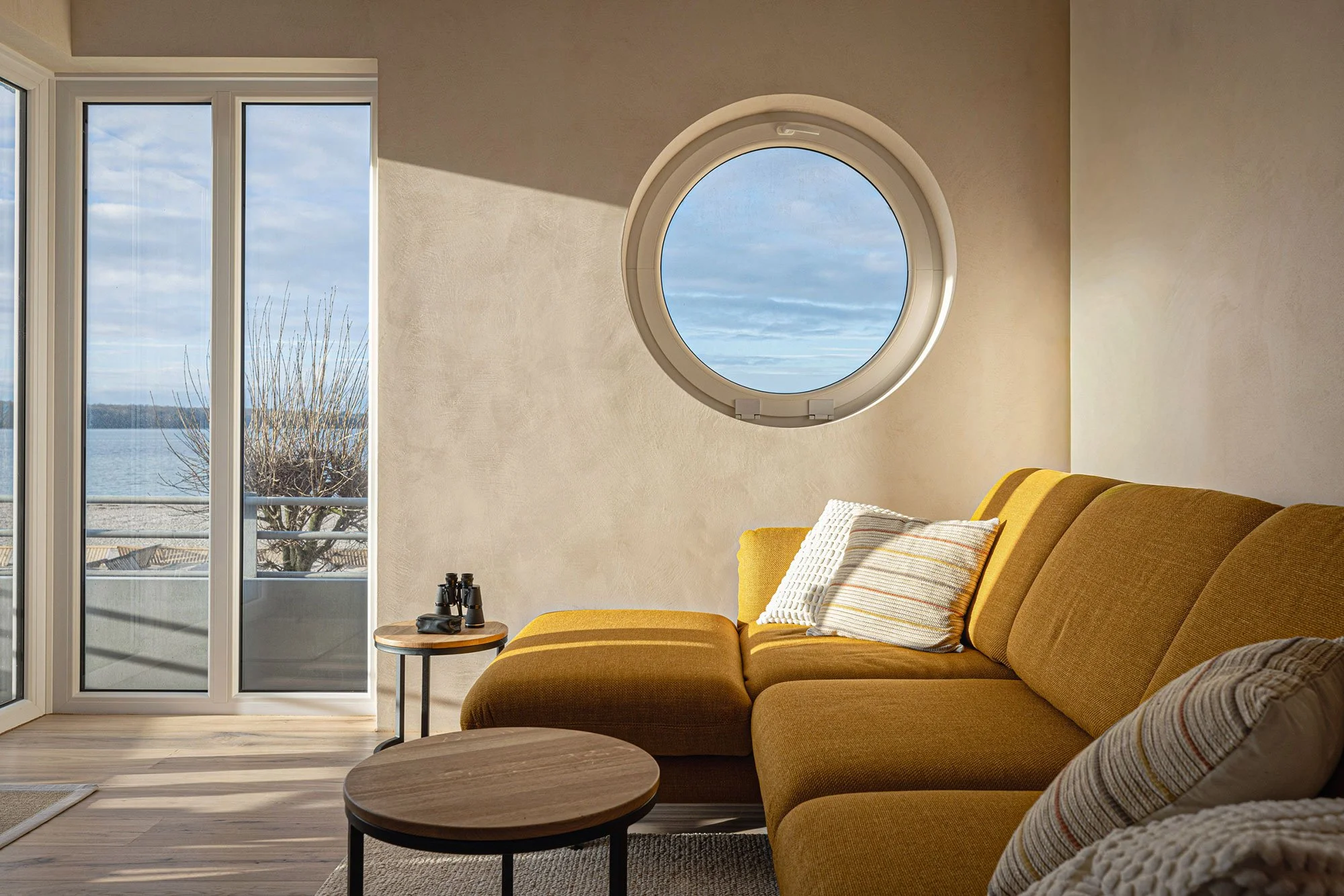 Wohnzimmer mit gelbem Sofa, Fenstern mit Blick auf Wasser und Baum, runder Fenster an der Wand, Kaffeetisch, kleine Vogelbeobachtungsoptik auf einem Beistelltisch.