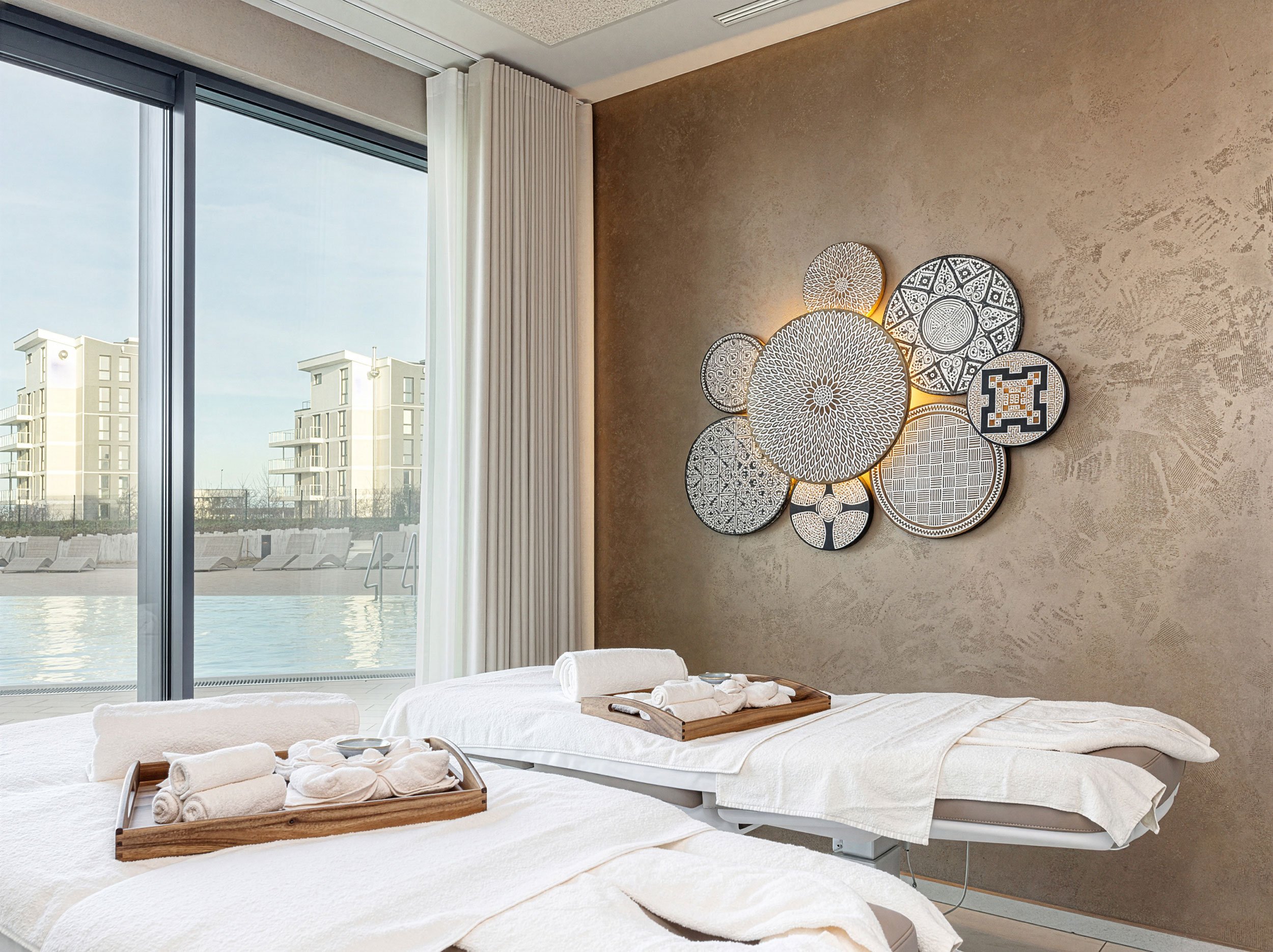 Ein Spa-Zimmer mit zwei Massagetischen, weißen Handtüchern, einem Blick auf einen Pool im Freien und moderne Wanddekoration