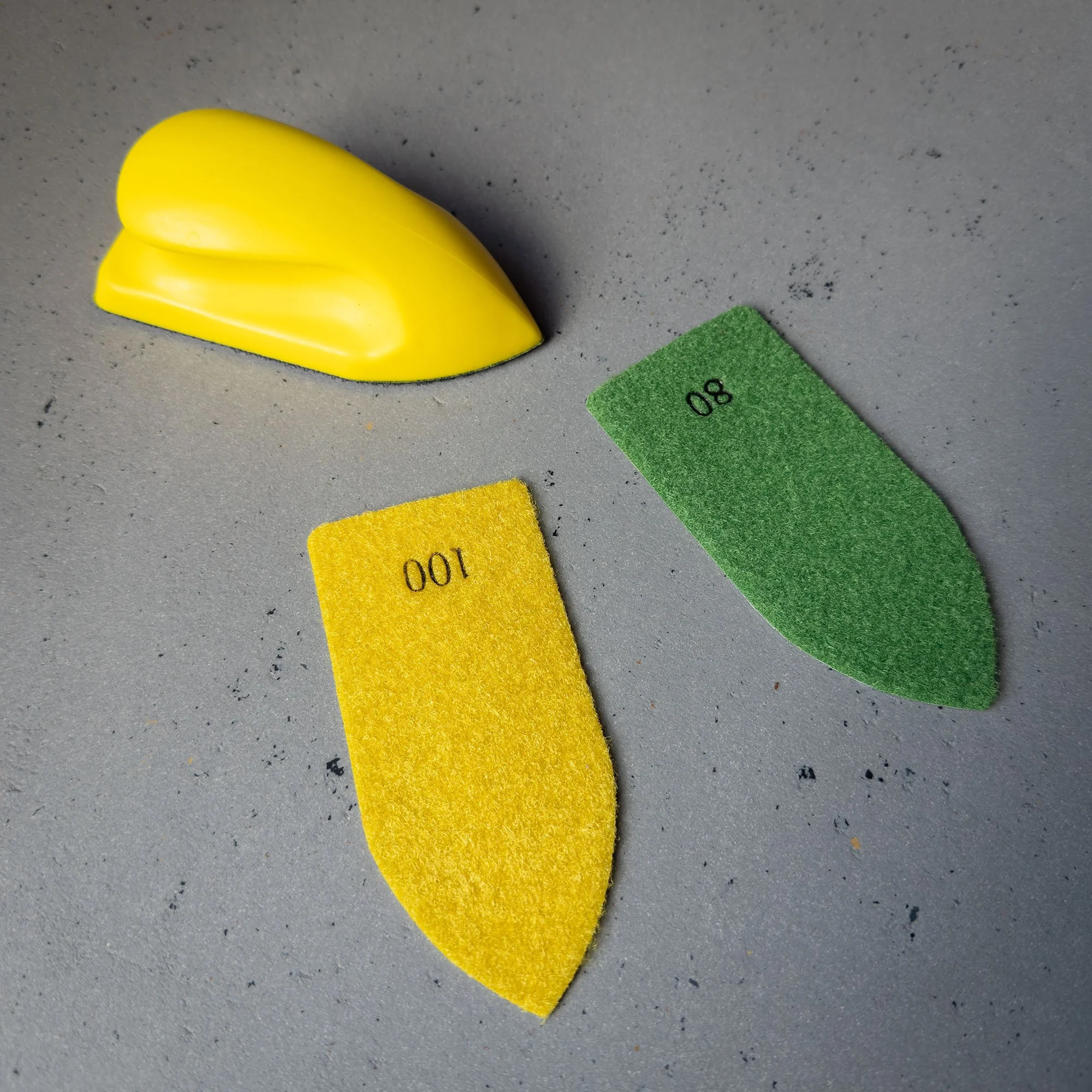 Kunststoffstück in Form eines Zitronens, zwei Schleifpads in Gelb und Grün, auf grauem Hintergrund.