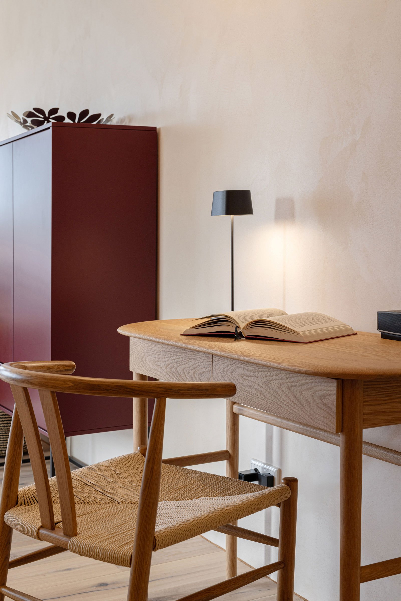 Ein Holztisch mit einem offenen Buch und einem Lautsprecher, im Hintergrund eine Stehlampe und ein rotes Schränkchen, Raum mit neutralen Wänden.