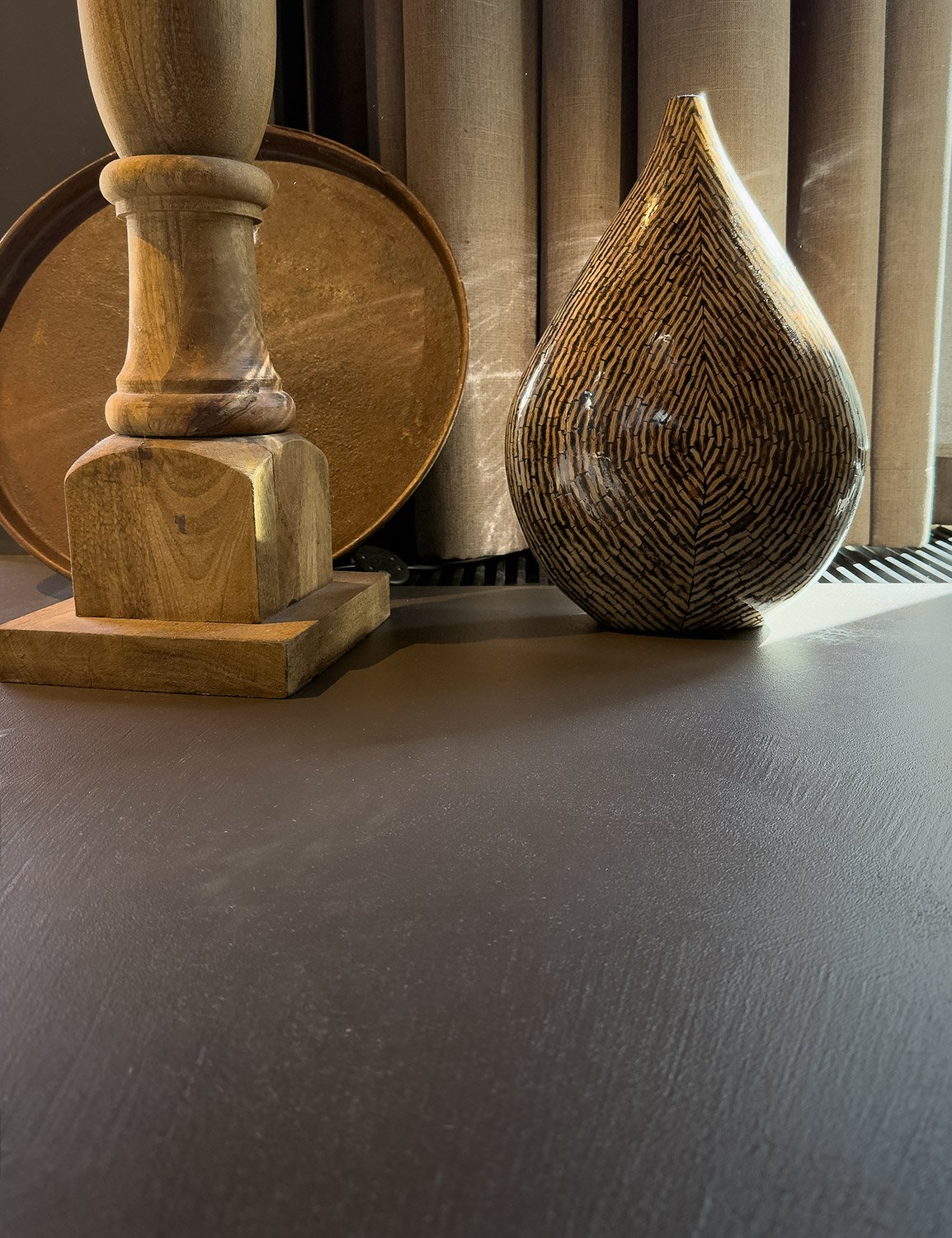 Holzdeko mit einer Holzpyramide, einem großen Pott und einer Holzvase mit Maserung auf einer Oberfläche vor einem Fenster mit beige Vorhängen.