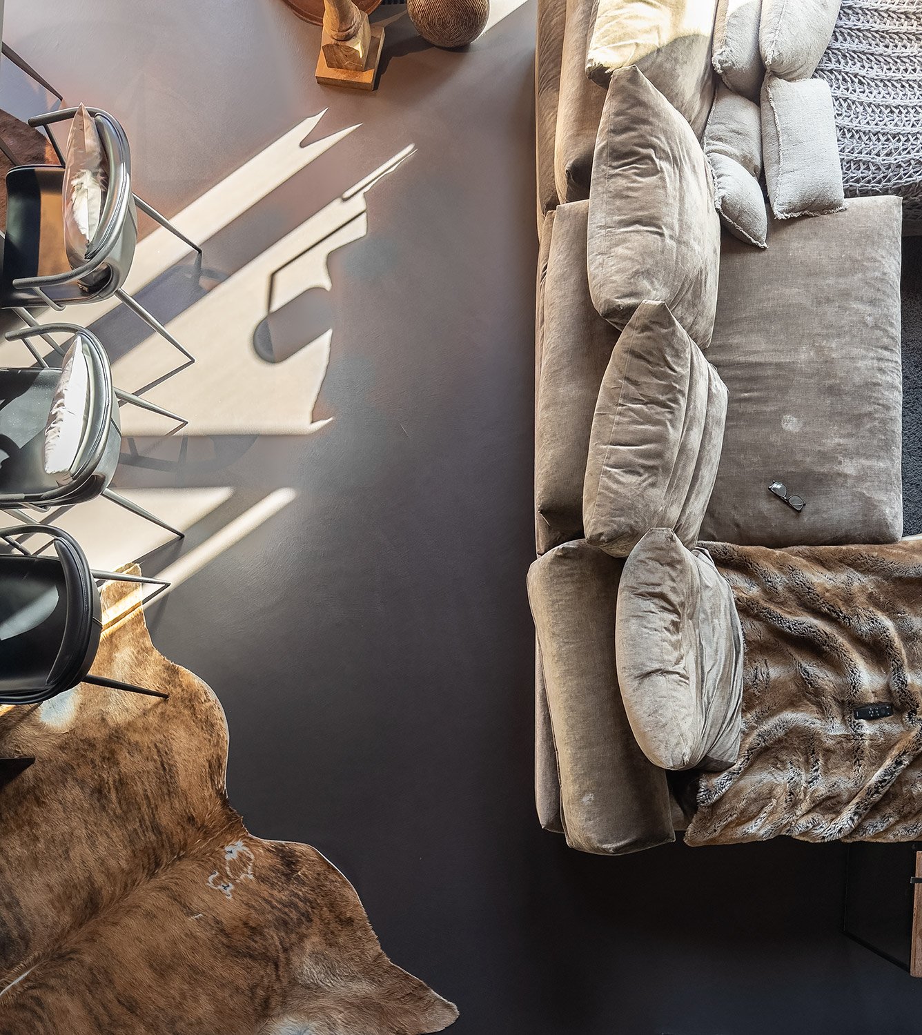 Ansicht eines Wohnzimmers mit einem grauen Sofa, mehreren Kissen, einer Brille auf dem Sofa, einem braunen Fellteppich, einem Esstisch mit Stühlen und Sonnenlicht, das Schatten an die Wand wirft.