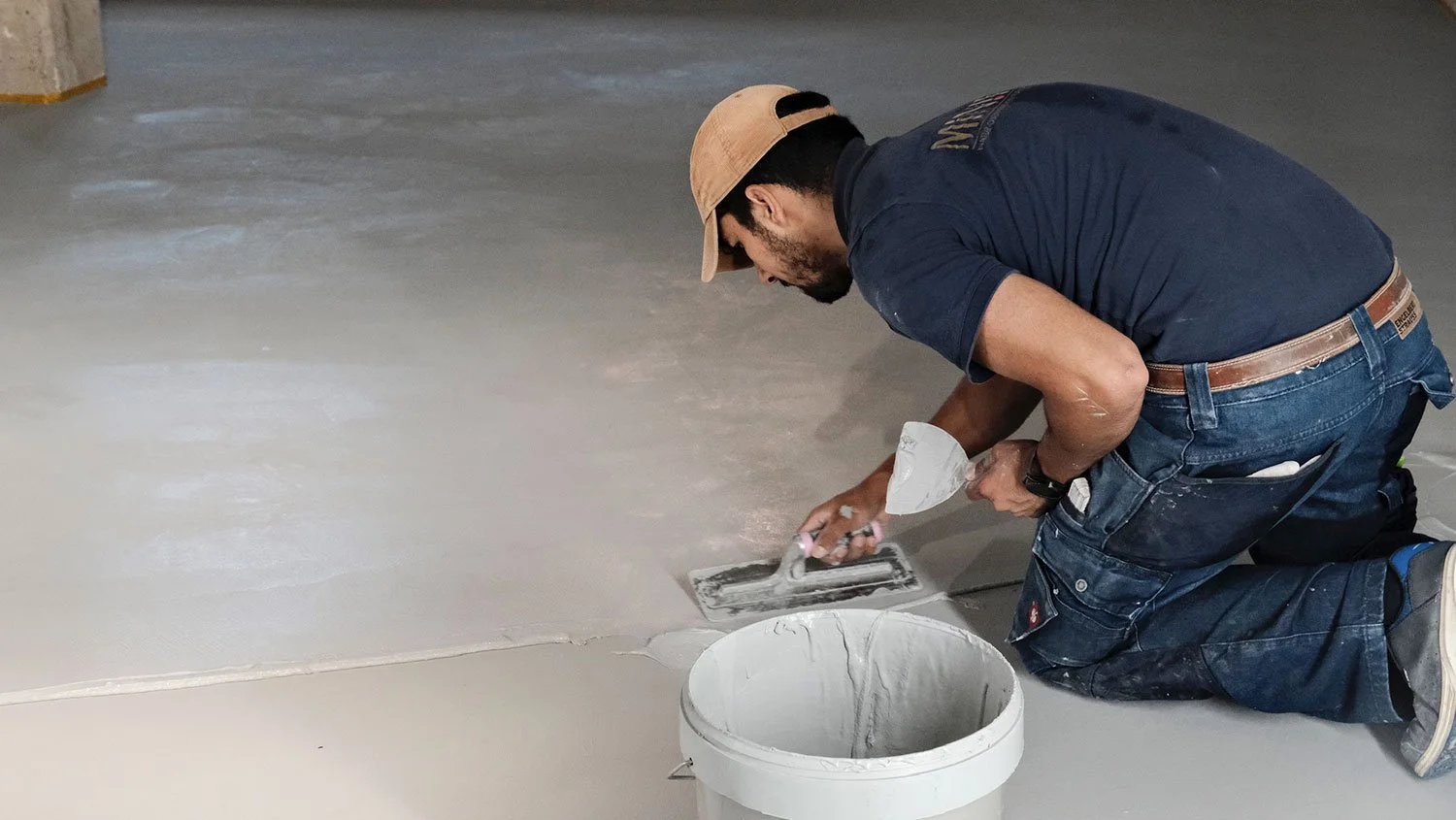 Ein Mann legt Fliesen auf den Boden, kniet dabei und benutzt ein Werkzeug, mit einem Eimer voller Mörtel neben ihm.