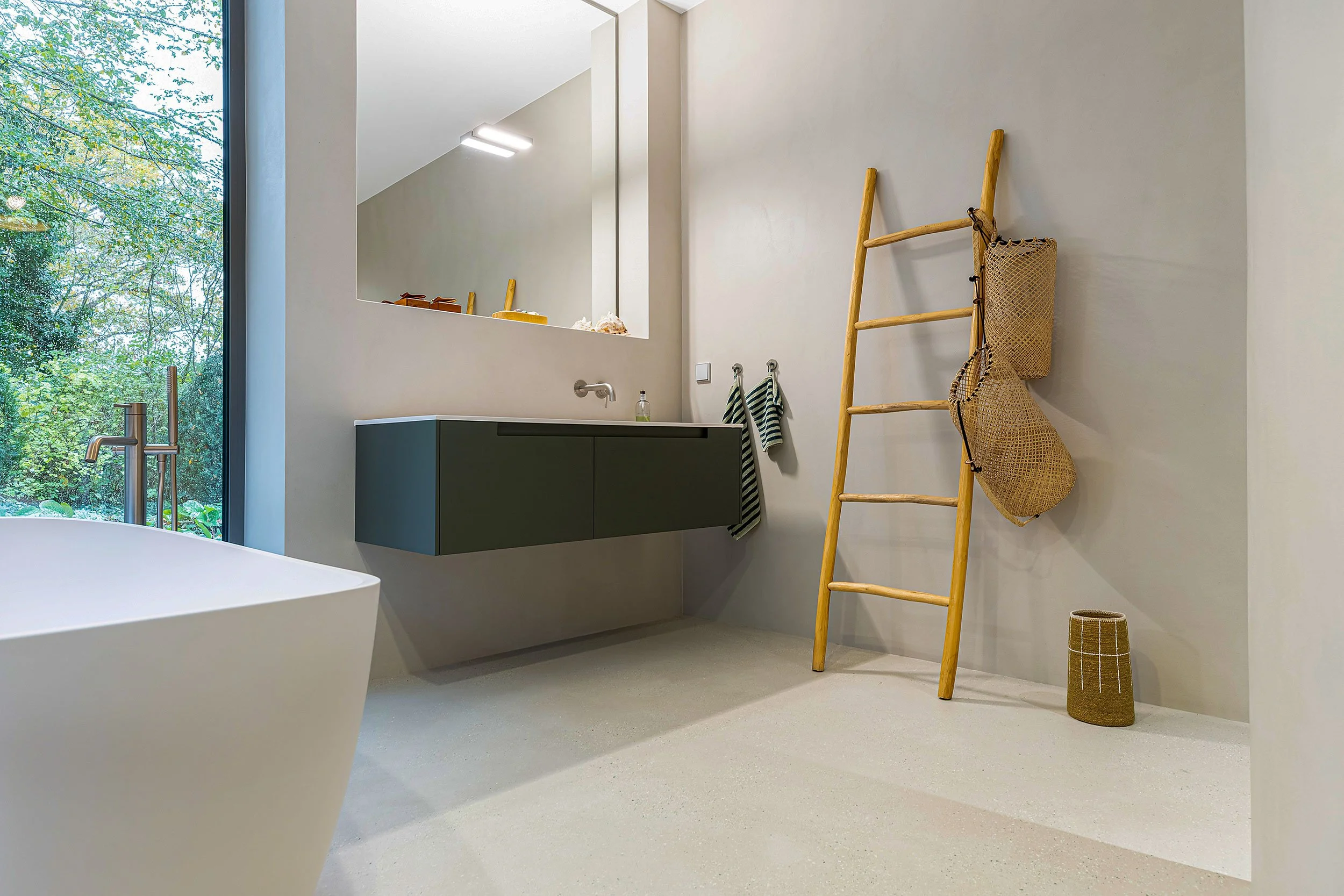 Ein minimalistisches Badezimmer mit einer freistehenden Badewanne, einem schwarzen Waschtischunizio mit Spiegel, einem Wandhaken mit Handtüchern, einer Holzleiter mit Körben und einem großen Fenster mit Blick auf grüne Bäume.