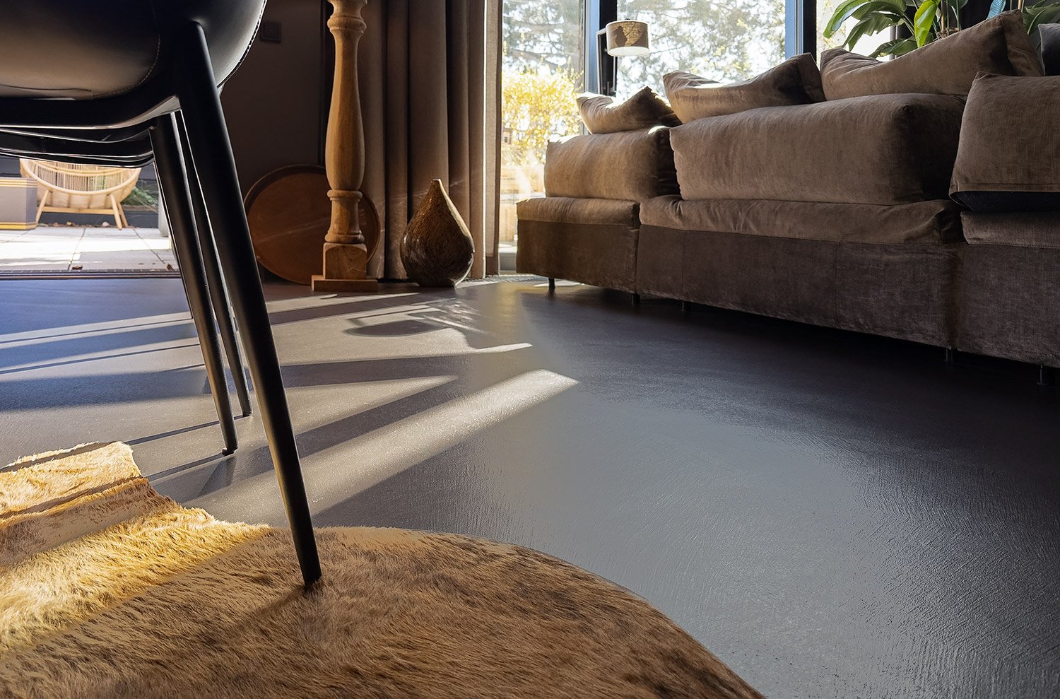 Wohnzimmer mit braunem Sofa, dunklem Boden, einem Stuhl, Teppich und Dekorationsgegenständen nahe dem Fenster, durch das Sonnenlicht einfällt.