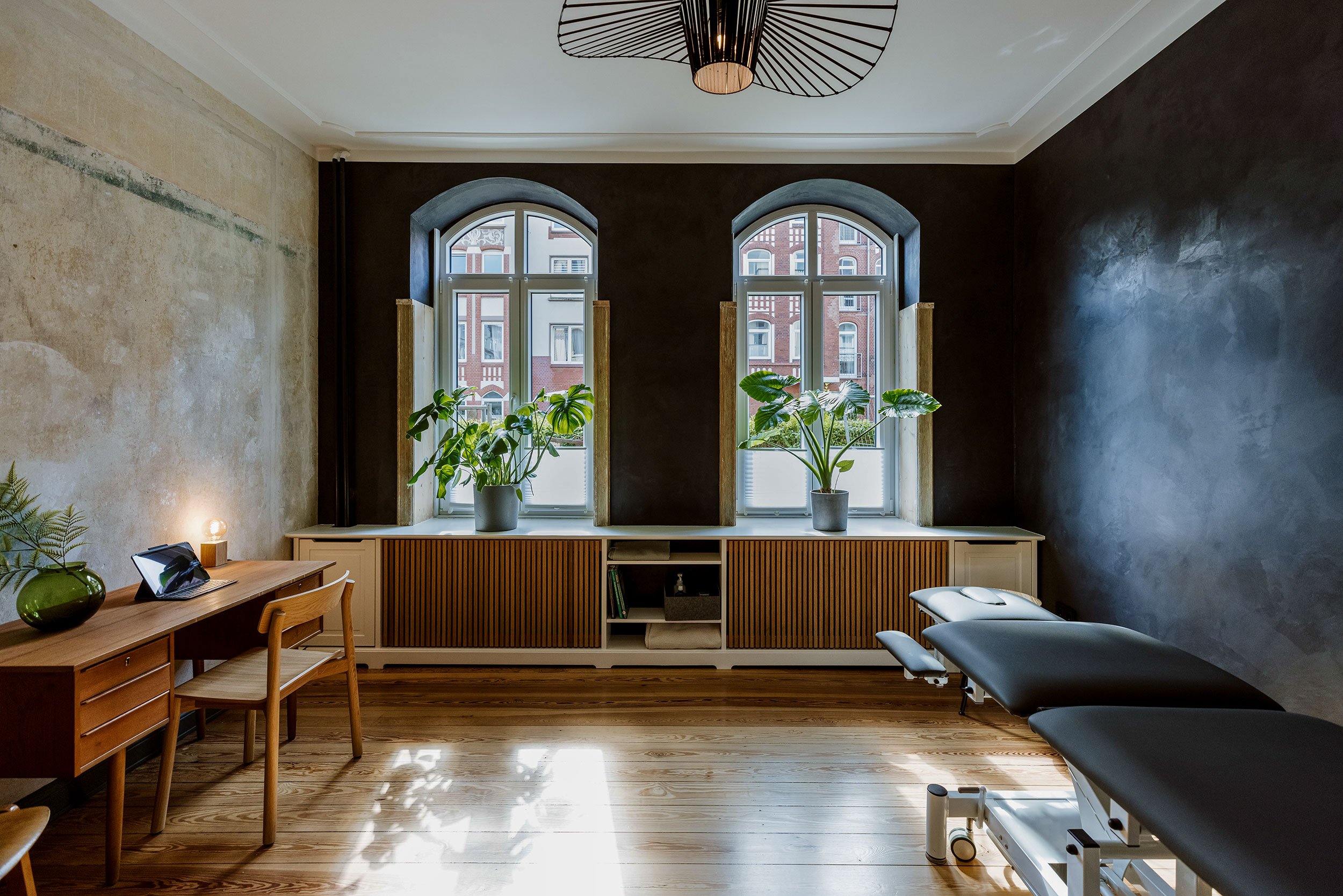 Behandlungszimmer mit zwei großen Fenster, Pflanzen auf der Fensterbank, Holztisch, Stuhl, dunkel gestrichene Wand, Behandlungsliege, natürliche Lichtquelle.