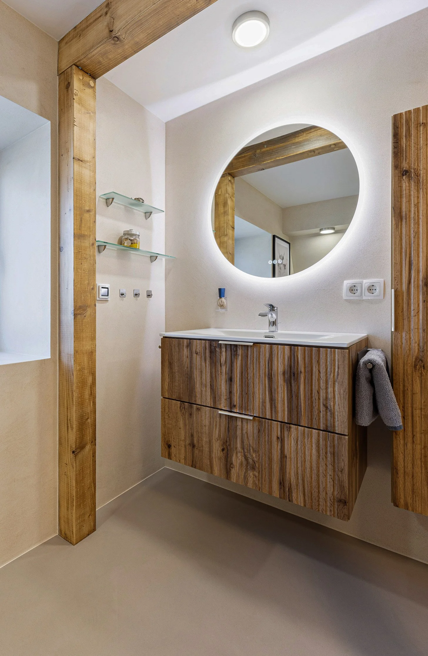 Moderner Badezimmerarmaturen mit Holzdesign, runder beleuchteter Spiegel und grauem Handtuch.