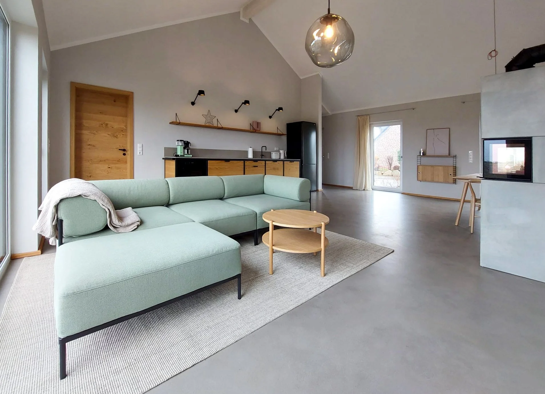 Offene, moderne Wohnzimmer- und Kitchenette im minimalistischen Stil mit grauem Boden, hellgrauen Wänden, schwarzem und Holz-Küchenschrank, grünem Sofa, zwei runden Holztischen, großen Fenstern und einem Kamin.