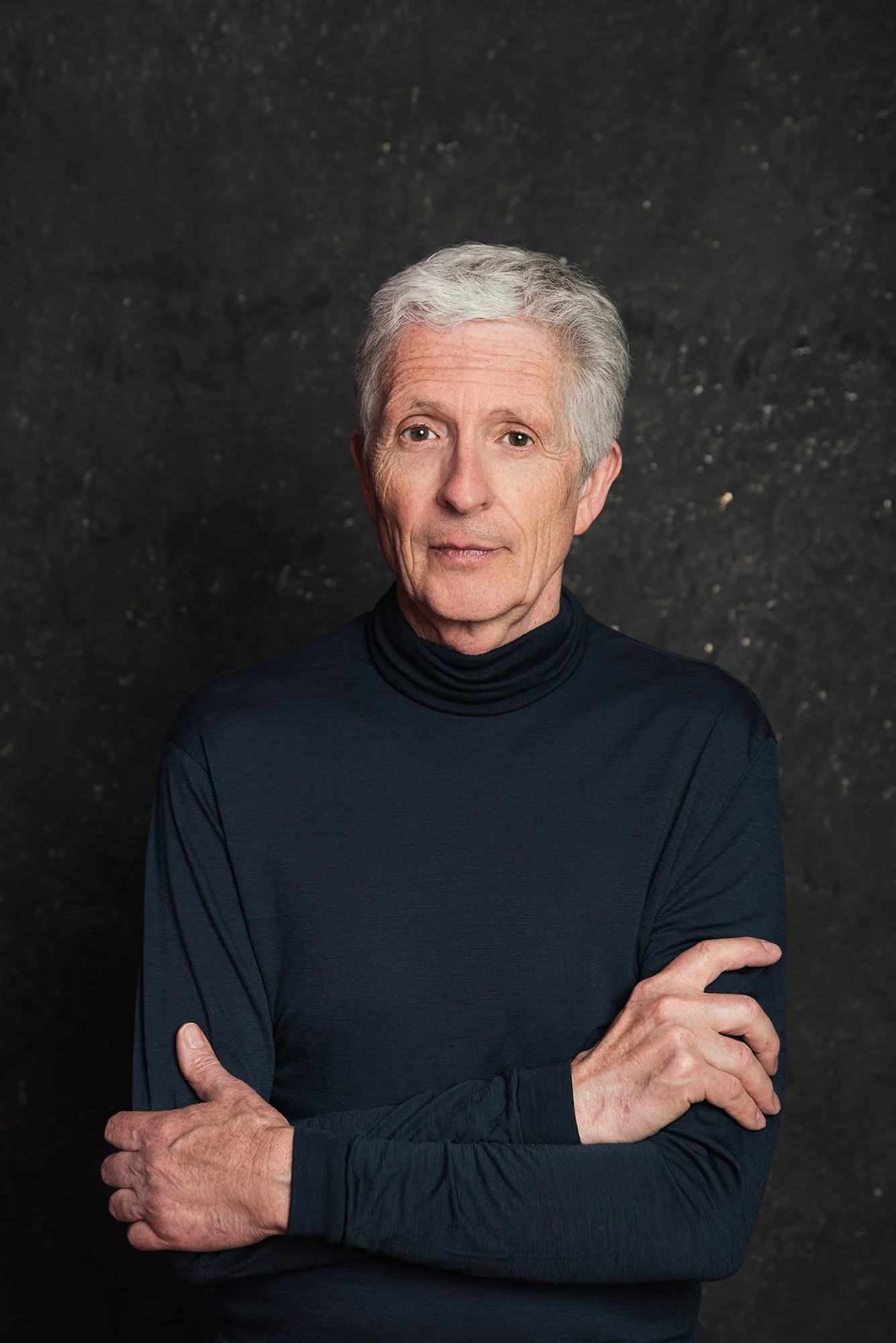 Ein älterer Mann mit grauem Haar trägt einen schwarzen Rollkragenpulli vor einem dunklen Hintergrund.