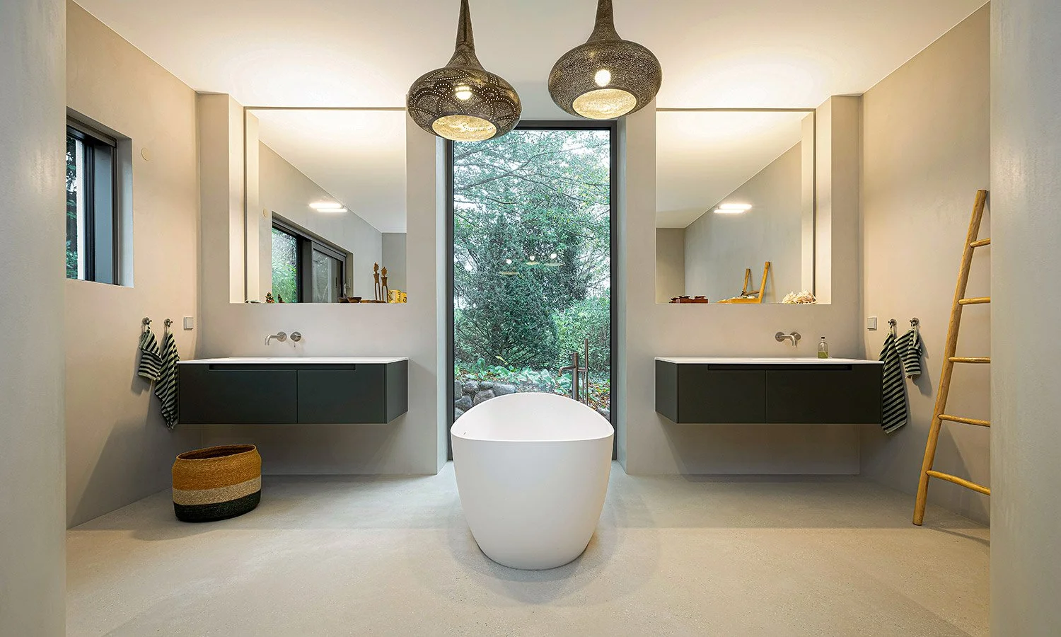 Modernes Badezimmer mit freistehender Badewanne vor großer Fensterfront, zwei Waschtische mit Spiegeln, grauen Unterschränken, Lichter, Dekorationsgegenständen und Handtüchern, Holzleiter, Korb, Blick ins Grüne draußen.