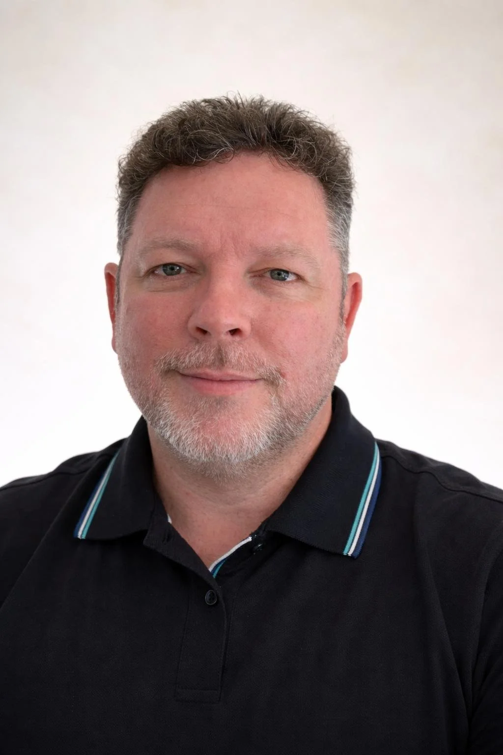 Ein Mann mit grauem Bart und blauen Augen trägt ein schwarzes Polo-Shirt vor einem weißen Hintergrund.