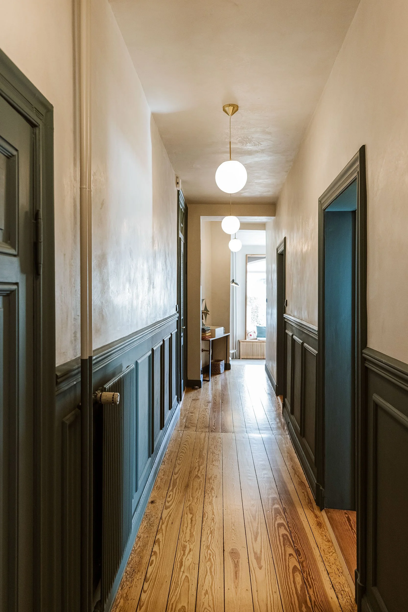 Langer Flur in einer klassischen Wohnung mit Tarkett-Holzboden, weißen Wänden, dunklen Wandpaneelen und sphericalen weißen Deckenleuchten, die den Flur beleuchten.