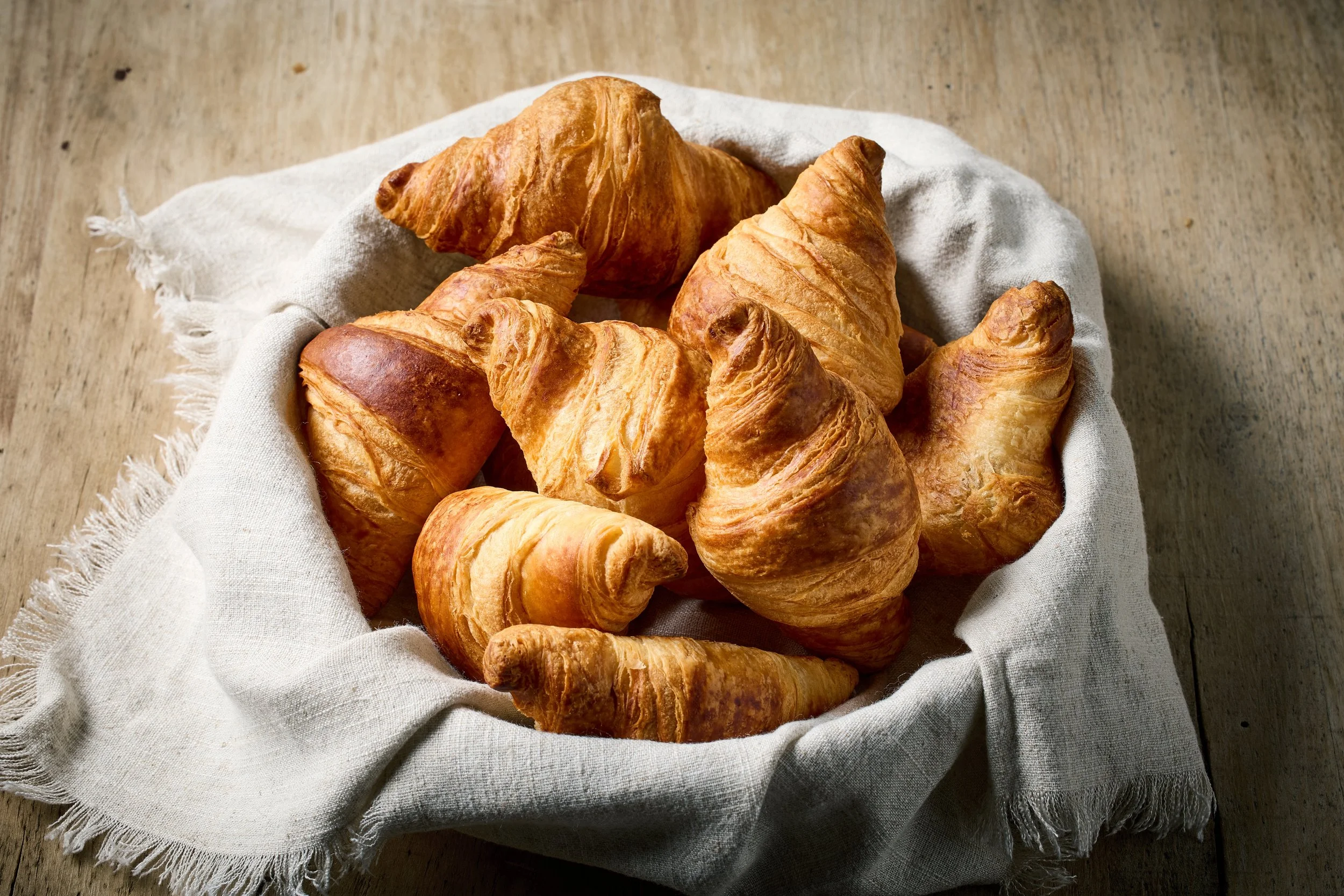A basket of golden-brown croissants in a white cloth basket on a wooden surface.