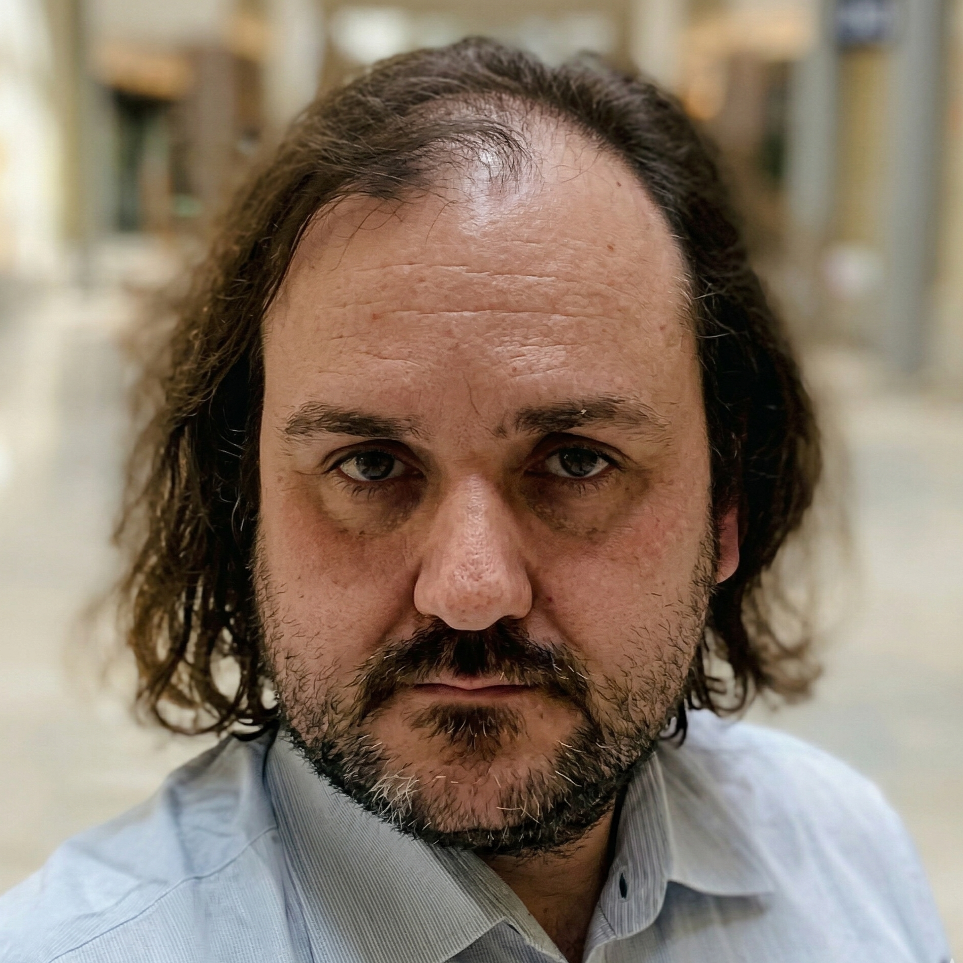 A close-up portrait of a man with shoulder-length wavy brown hair, a beard, and wearing a light blue collared shirt.