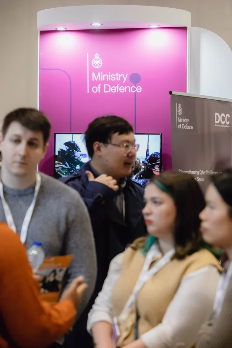 People gathered at a Ministry of Defence booth with pink background and displays, discussing cybersecurity topics.