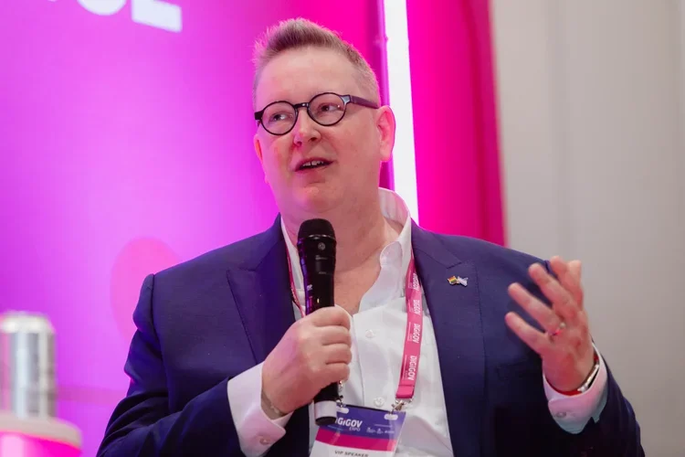 A person wearing glasses and a dark blue suit jacket speaking into a handheld microphone during an event, with a pink backdrop and a name badge hanging around their neck.