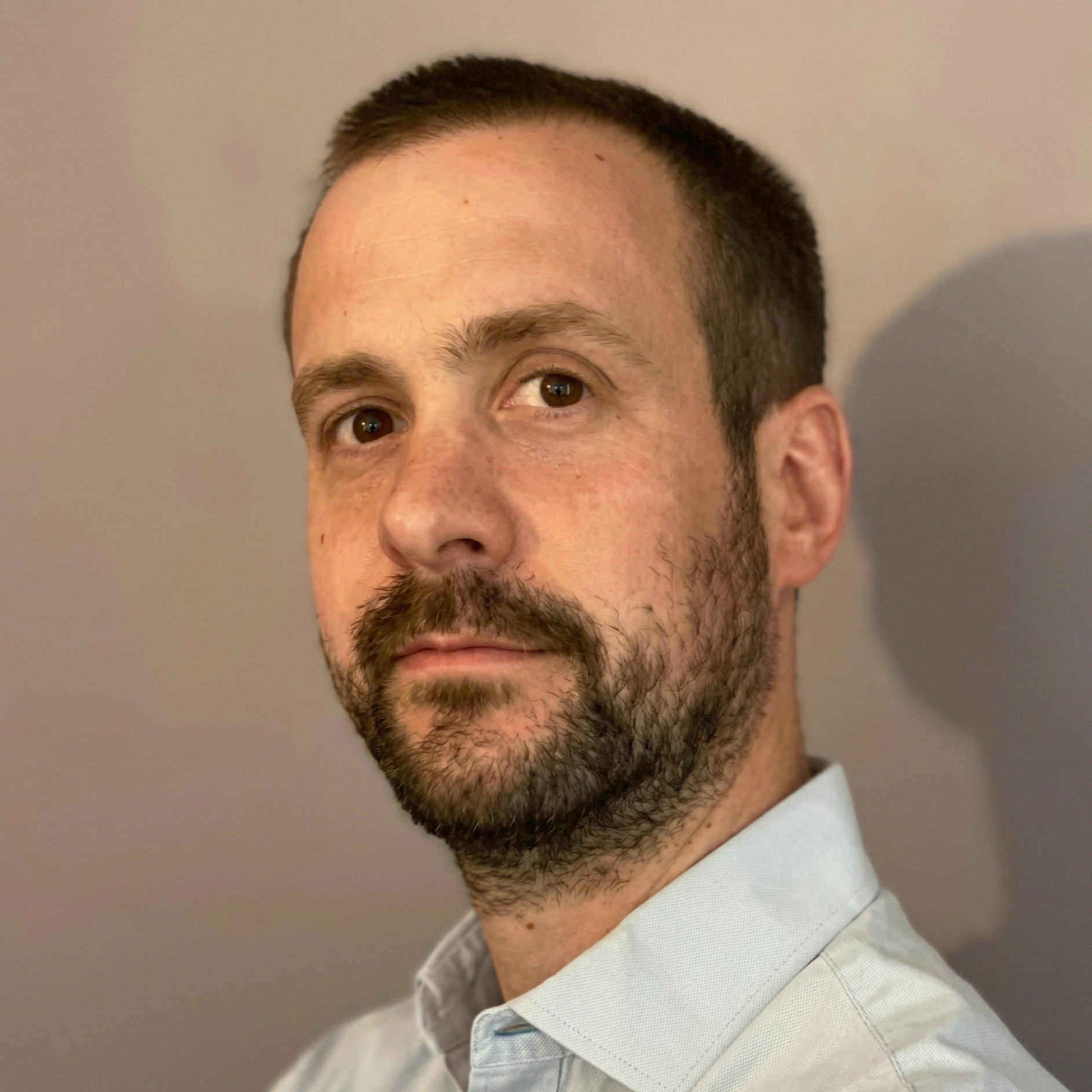 Close-up of a man with short dark hair, brown eyes, and a beard, wearing a light-colored collared shirt, standing against a plain beige wall, looking at the camera.