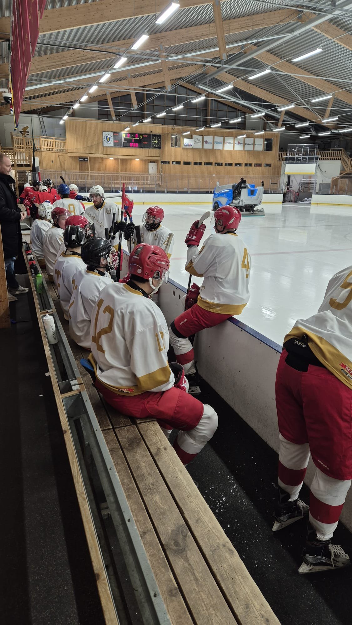 Hockeyspelare på bänken vid isen i en inomhusarena, några av dem med helmuts. En sopmaskin städar isen i bakgrunden under taket.
