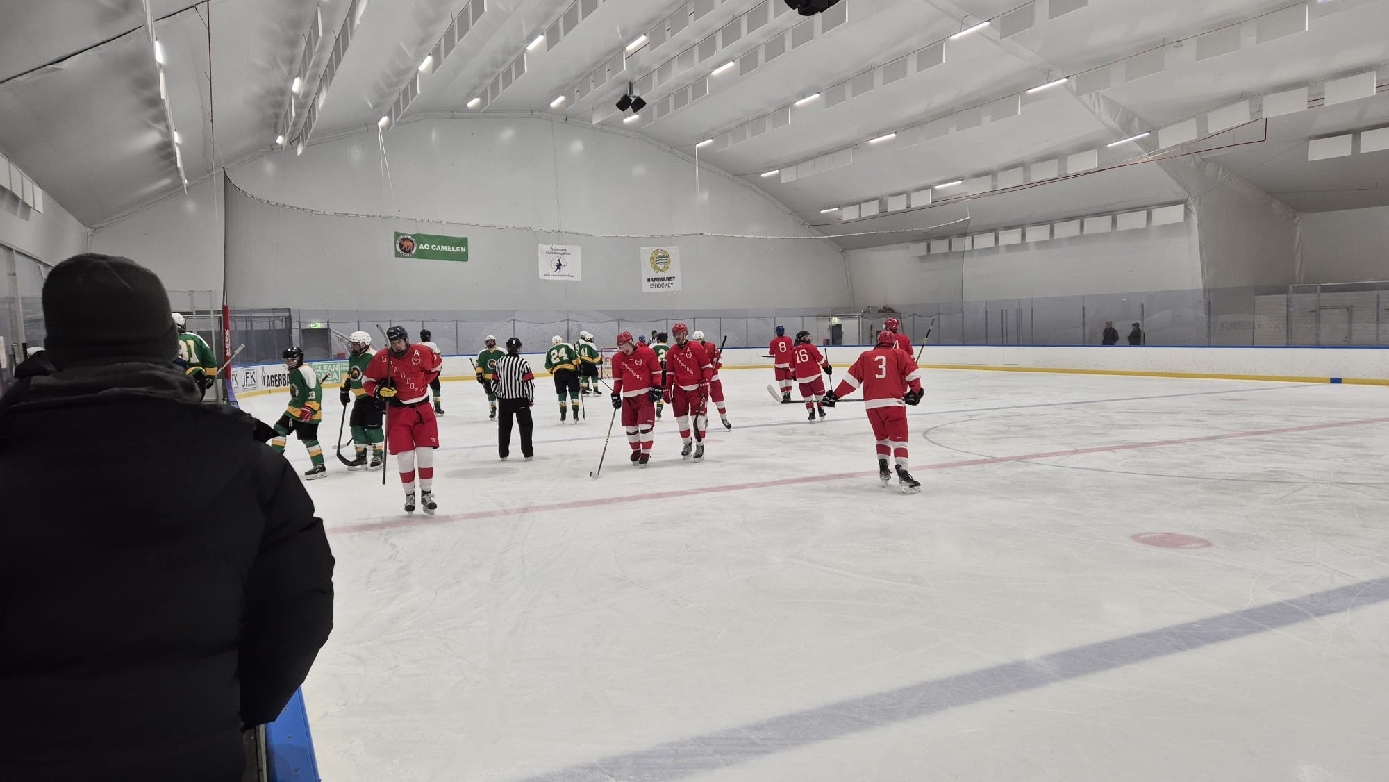 Hockeyspelare på isen efter en match, några går och andra står stilla, i en inomhusarena med vita väggar och taklampor.