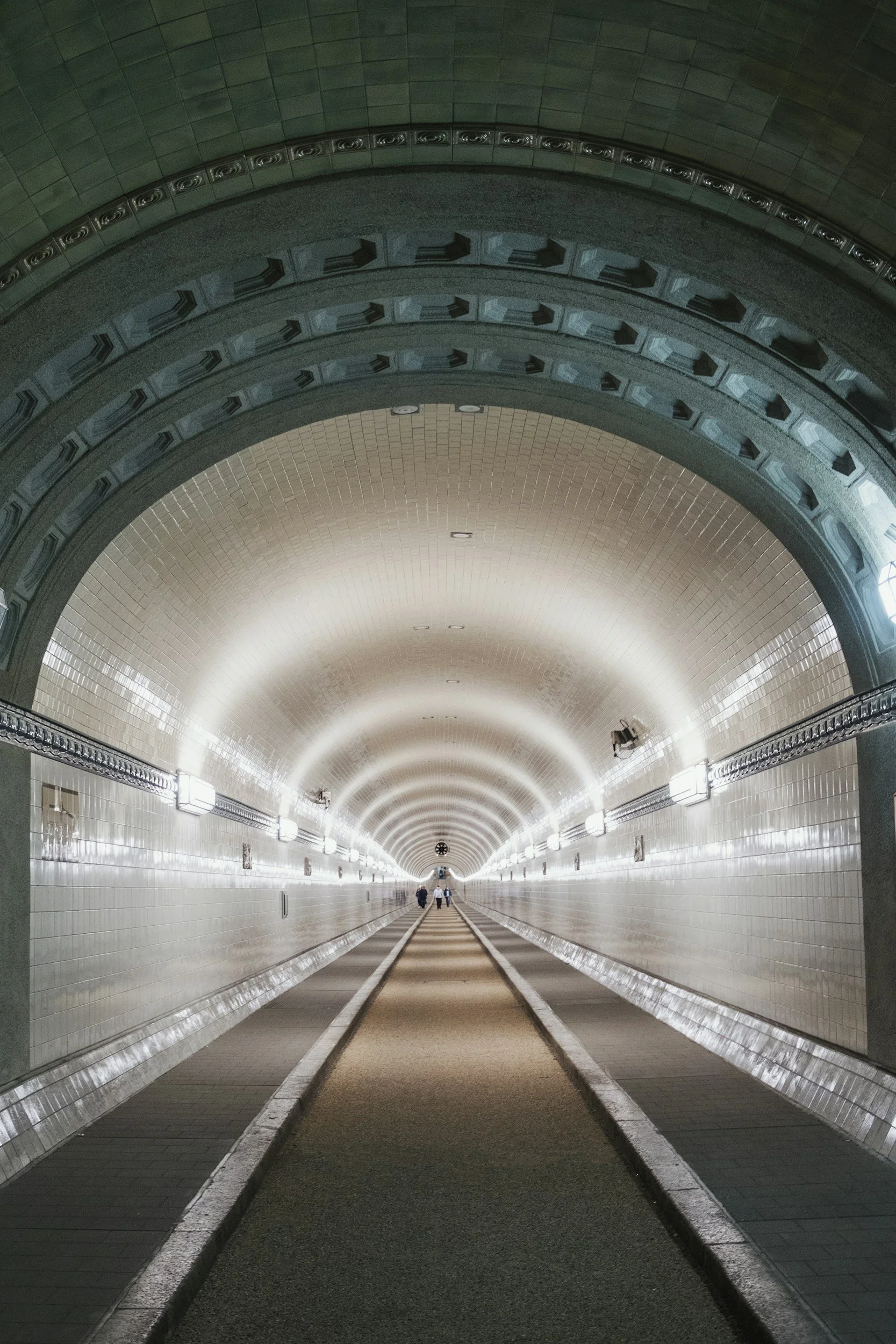 Langer Tunnel mit weißen Wandfliesen, Beleuchtung und Schienen auf dem Boden, mit zwei Personen, die am Ende des Tunnels gehen.