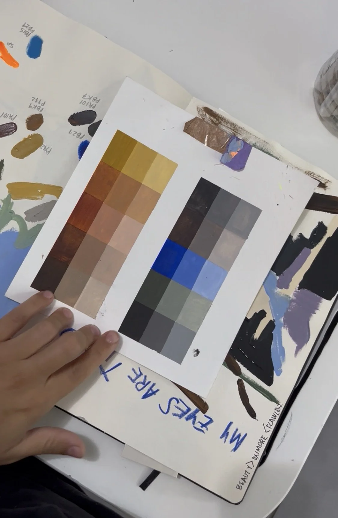 Close-up of a color palette comparison with swatches of various shades of brown, gray, blue, and other colors, alongside handwritten notes and a hand pointing at the palette. Artwork by © 2026 Aura Delamotte Barrera, All rights reserved.