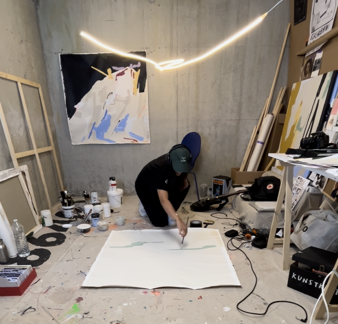 An artist in a studio working on a large canvas laid out on the floor, with various art supplies and tools scattered around, artwork on the walls, and a neon light shaped like a circle overhead. Artwork by © 2026 Aura Delamotte Barrera, All rights re