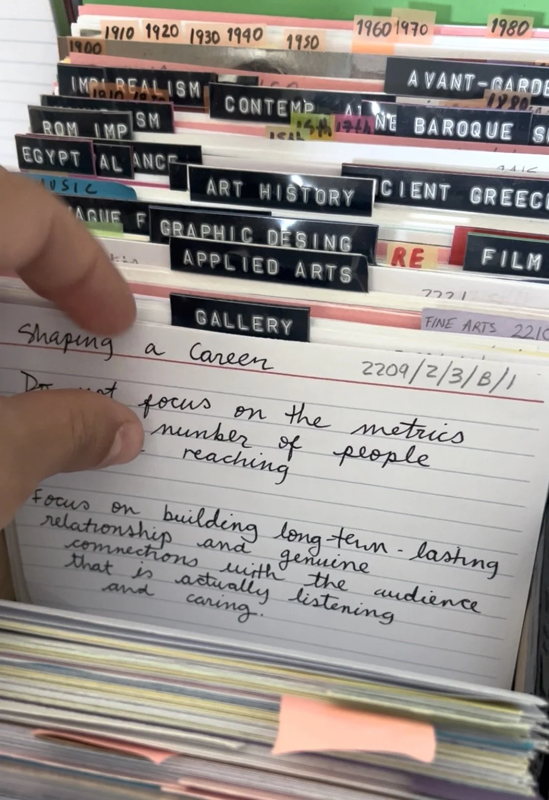 Close-up of handwritten notes on a page in a notebook, with a person's finger pointing at the text. The background shows organized tabs with labels for different history and art topics, including time periods and art styles, in a filing or storage system.