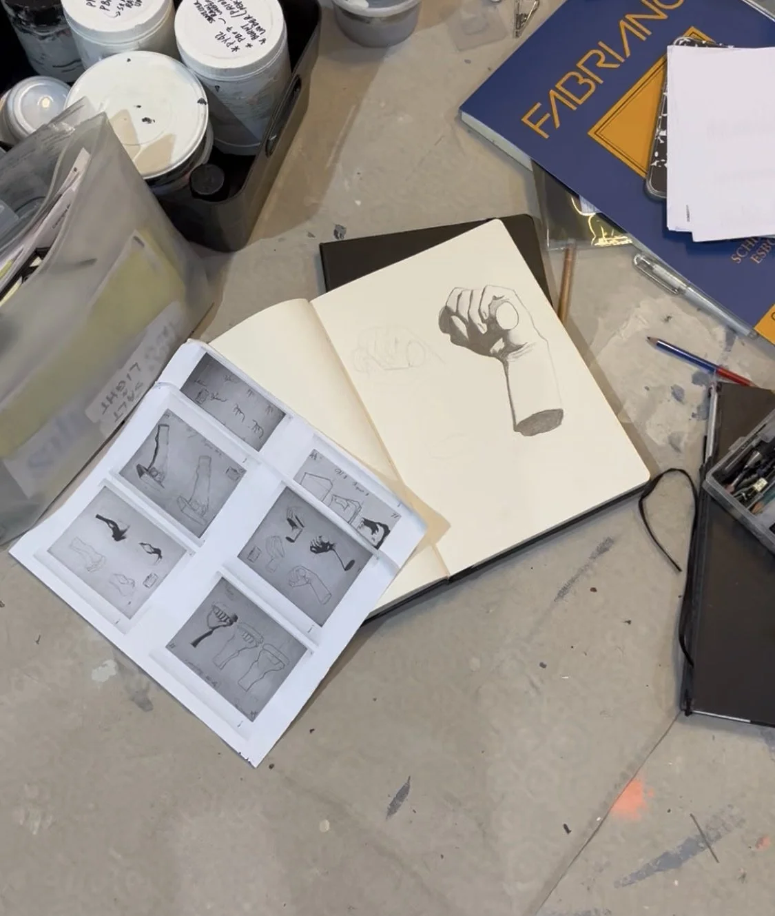A sketchbook with a drawing of a clenched fist on an open page, surrounded by art supplies, notebooks, and printed sketches on a cluttered table. Artwork by © 2026 Aura Delamotte Barrera, All rights reserved.