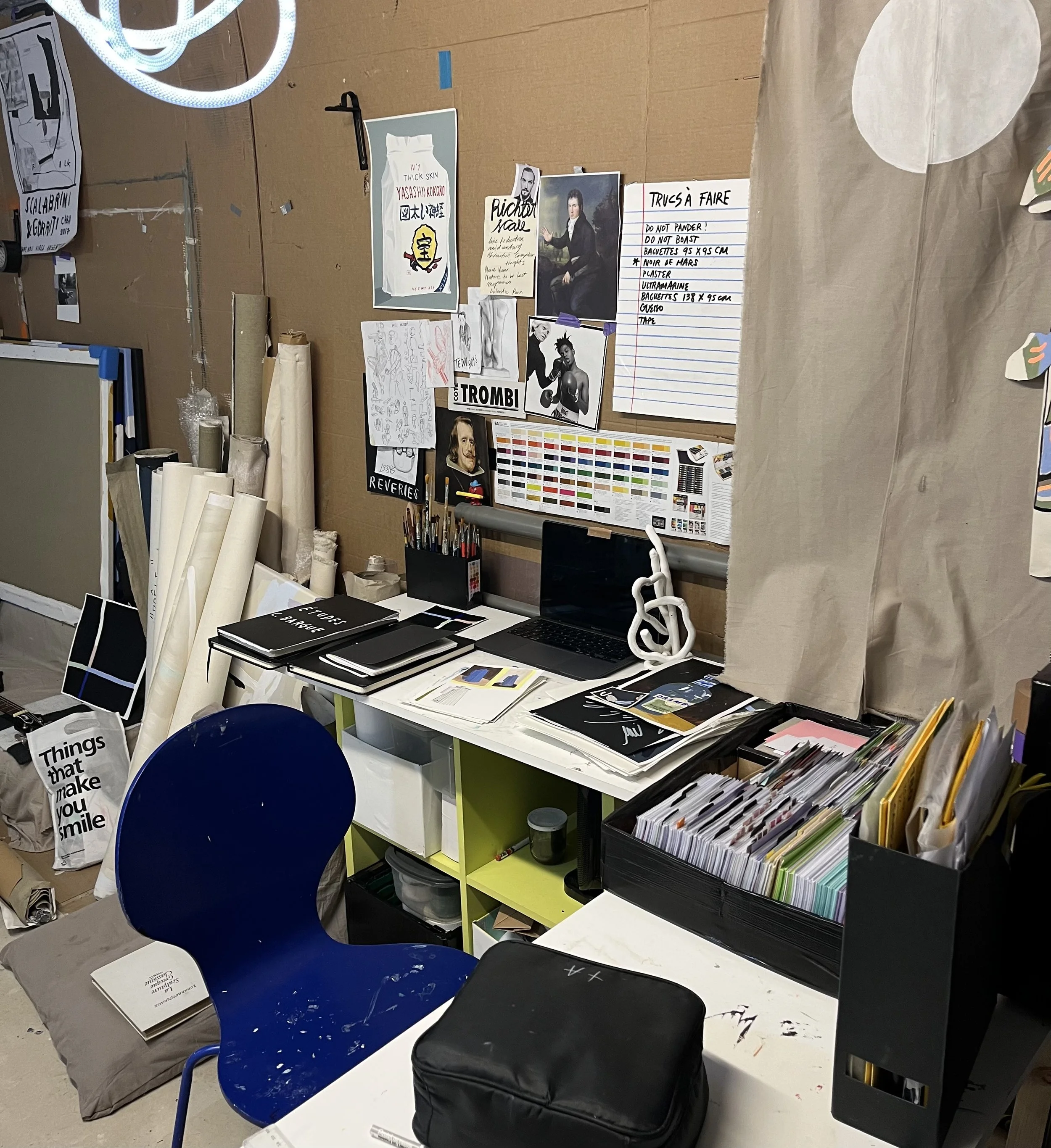 A cluttered workspace with a blue chair and a white desk containing a laptop, notebooks, and various office supplies. The background includes a wall covered with posters, sketches, and notes, with rolled-up papers leaning against it. There is a cubical shelf with organized folders
