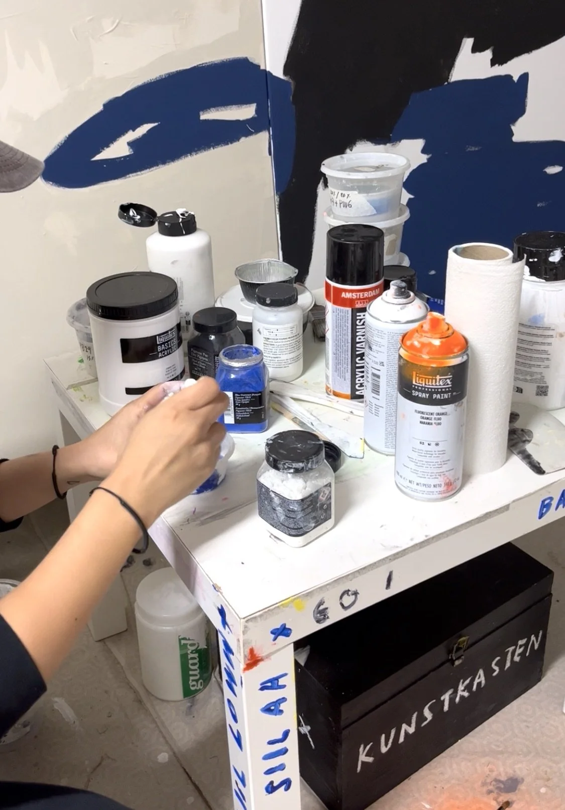 Art supplies including spray paint cans, jars, and bottles on a small, painted table with a mirror behind it. Artwork by © 2026 Aura Delamotte Barrera, All rights reserved.