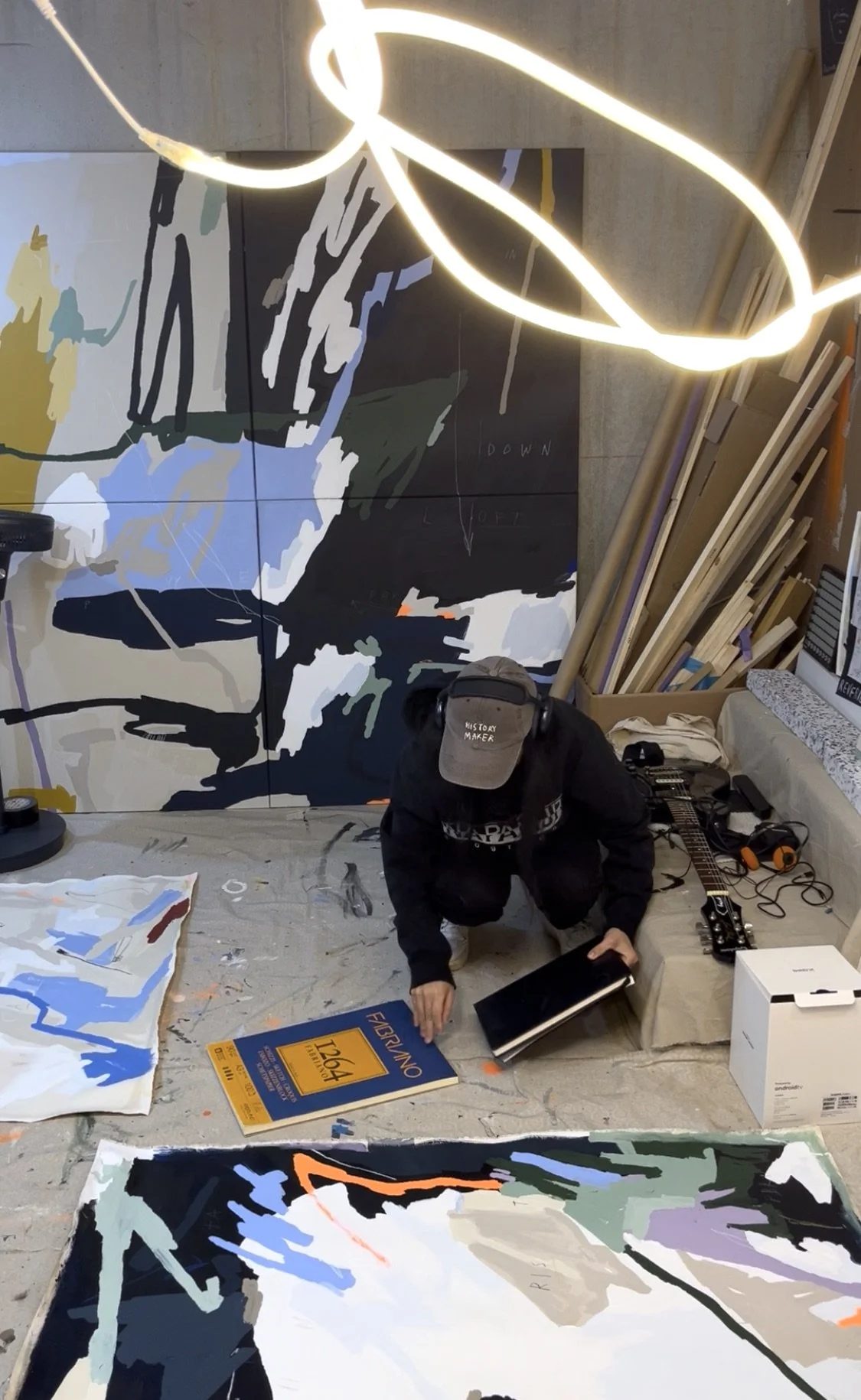An artist is working on a large abstract painting in a studio, surrounded by art supplies and materials, with a prominent glowing light fixture overhead. Artwork by © 2026 Aura Delamotte Barrera, All rights reserved.