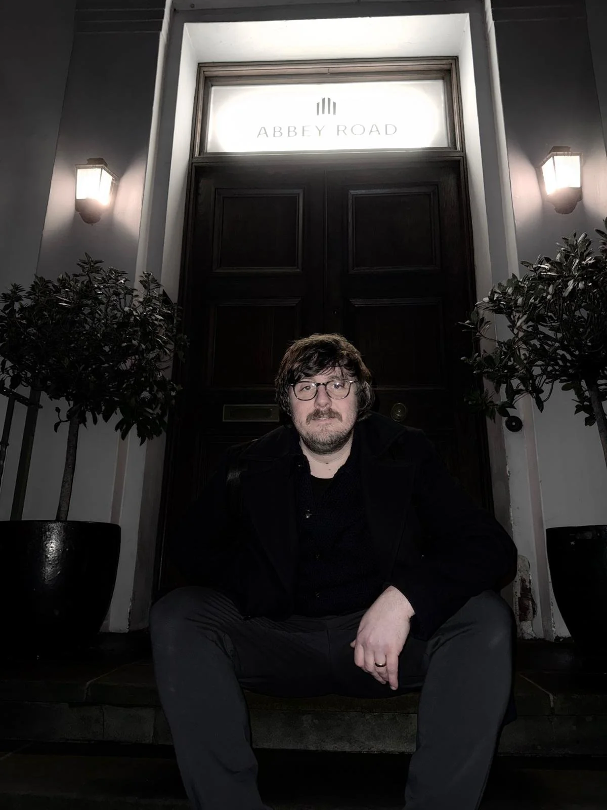 A man with glasses and facial hair sitting on porch steps in front of a dark wooden door with illuminated sign reading 'Abbey Road'. Two potted plants on either side of the steps and wall-mounted lights.