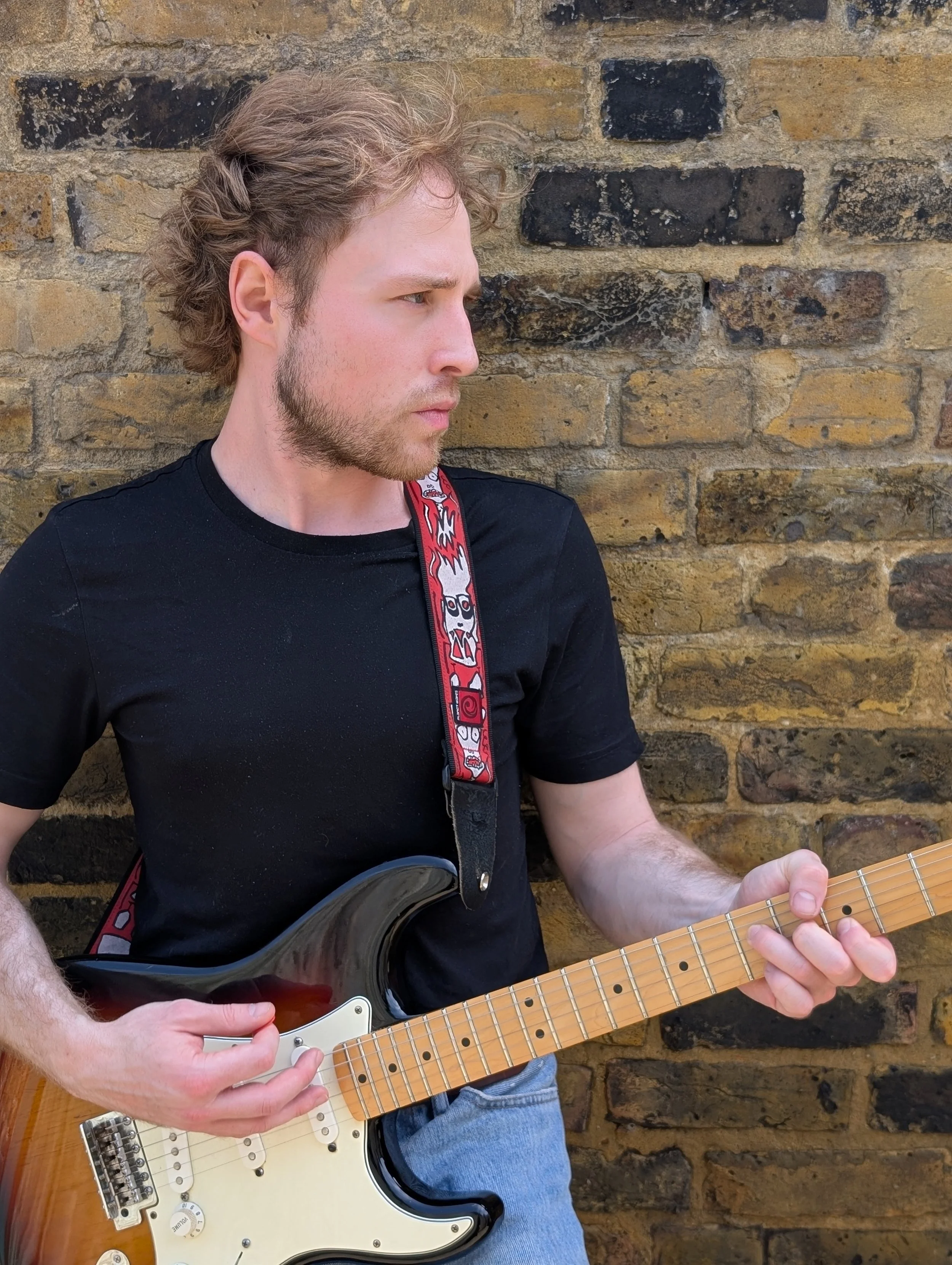 A man with light skin, brown curly hair, and a beard, wearing a black shirt, playing an electric guitar with a sunburst finish, against a brick wall background.
