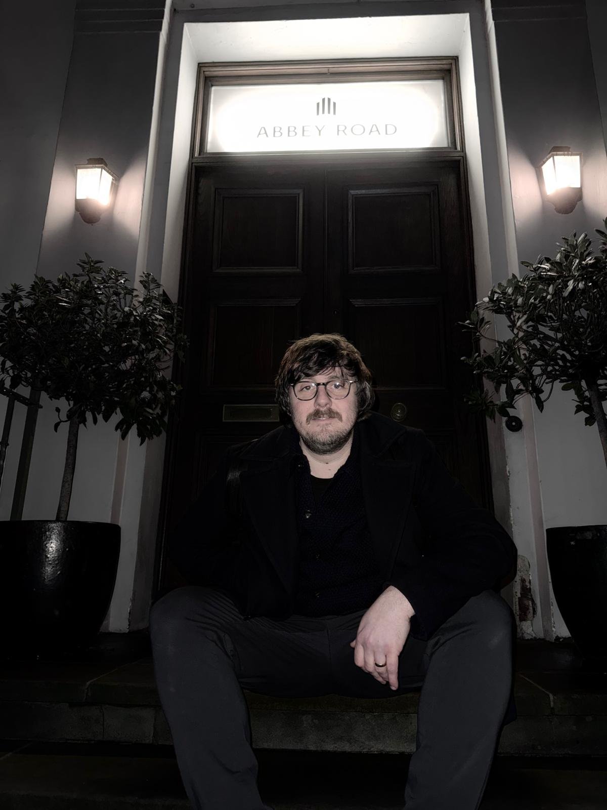 Man with glasses and a beard sitting in front of a large, dark wooden door with a sign above it that reads "Abbey Road". Two potted plants are on either side of the door, and there are wall-mounted lights on each side.