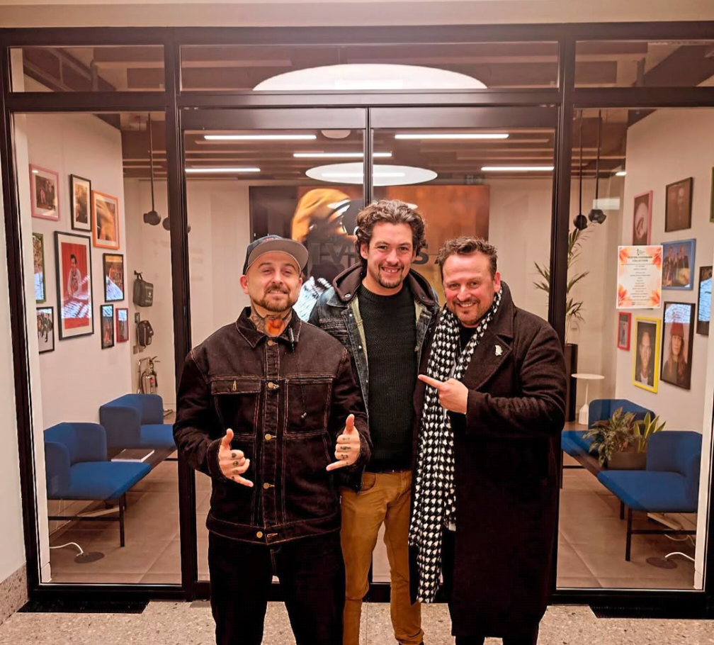 Three men standing inside a glass enclosed room, smiling and posing for a photo. The room has framed photos on the walls and blue chairs on the sides.