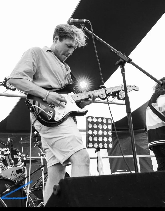 A young man playing an electric guitar on stage with a microphone in front of him, outdoor setting.