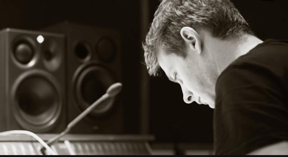 A man with short hair sitting in front of music speakers and a microphone in a recording studio.