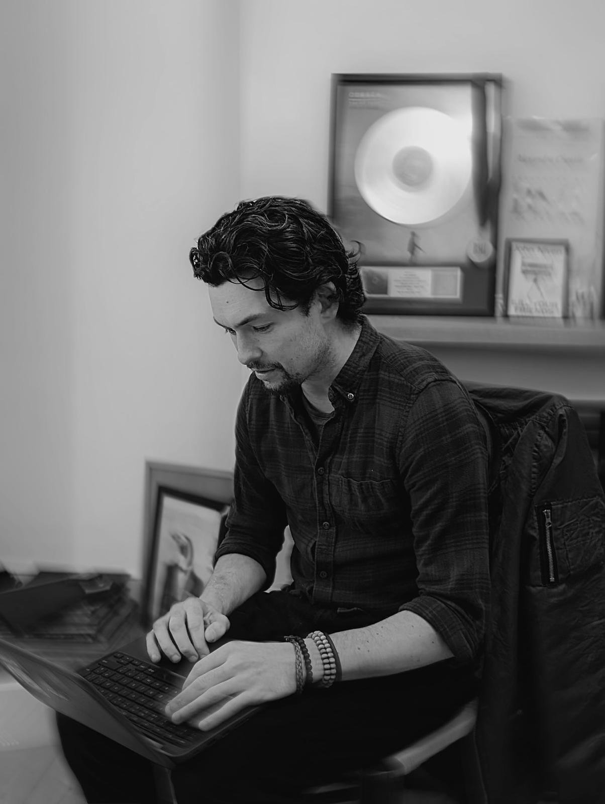 A man with dark wavy hair, wearing a dark checkered shirt and wristbands, sitting at a desk working on a laptop with a focused expression.
