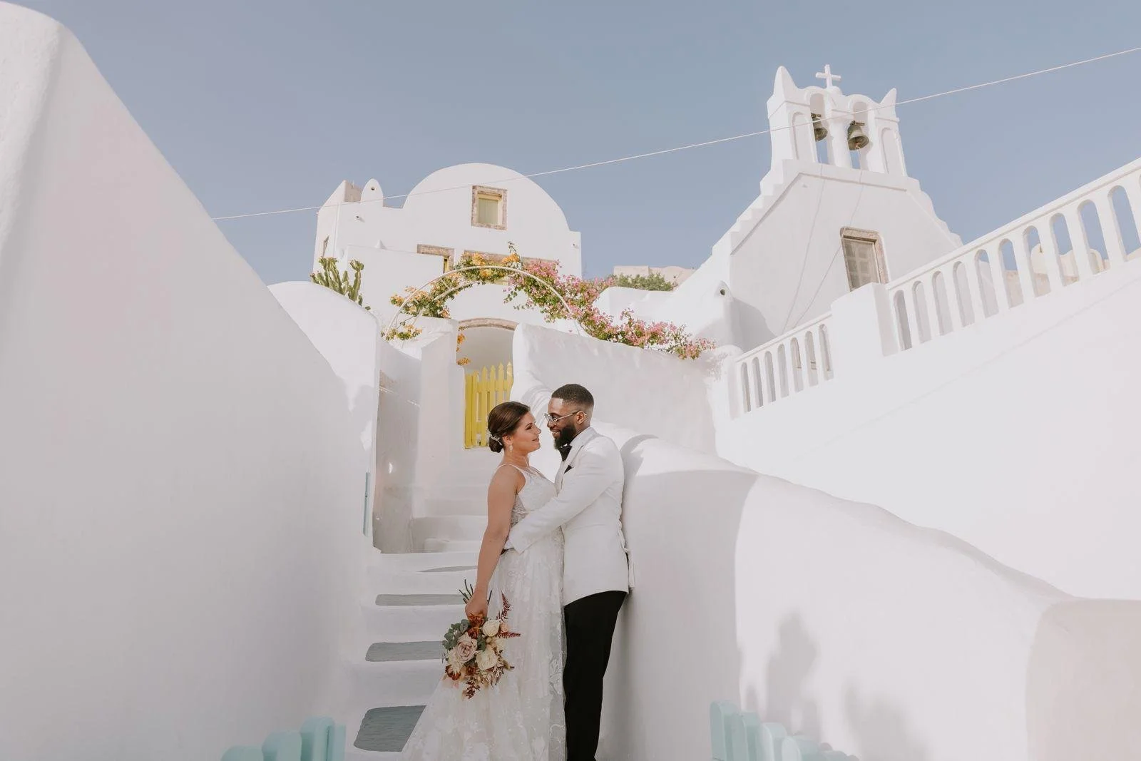 Un couple de mariés debout sur des escaliers blancs devant un bâtiment méditerranéen blanc avec des cloches et une croix, dans un cadre ensoleillé et bleu ciel.