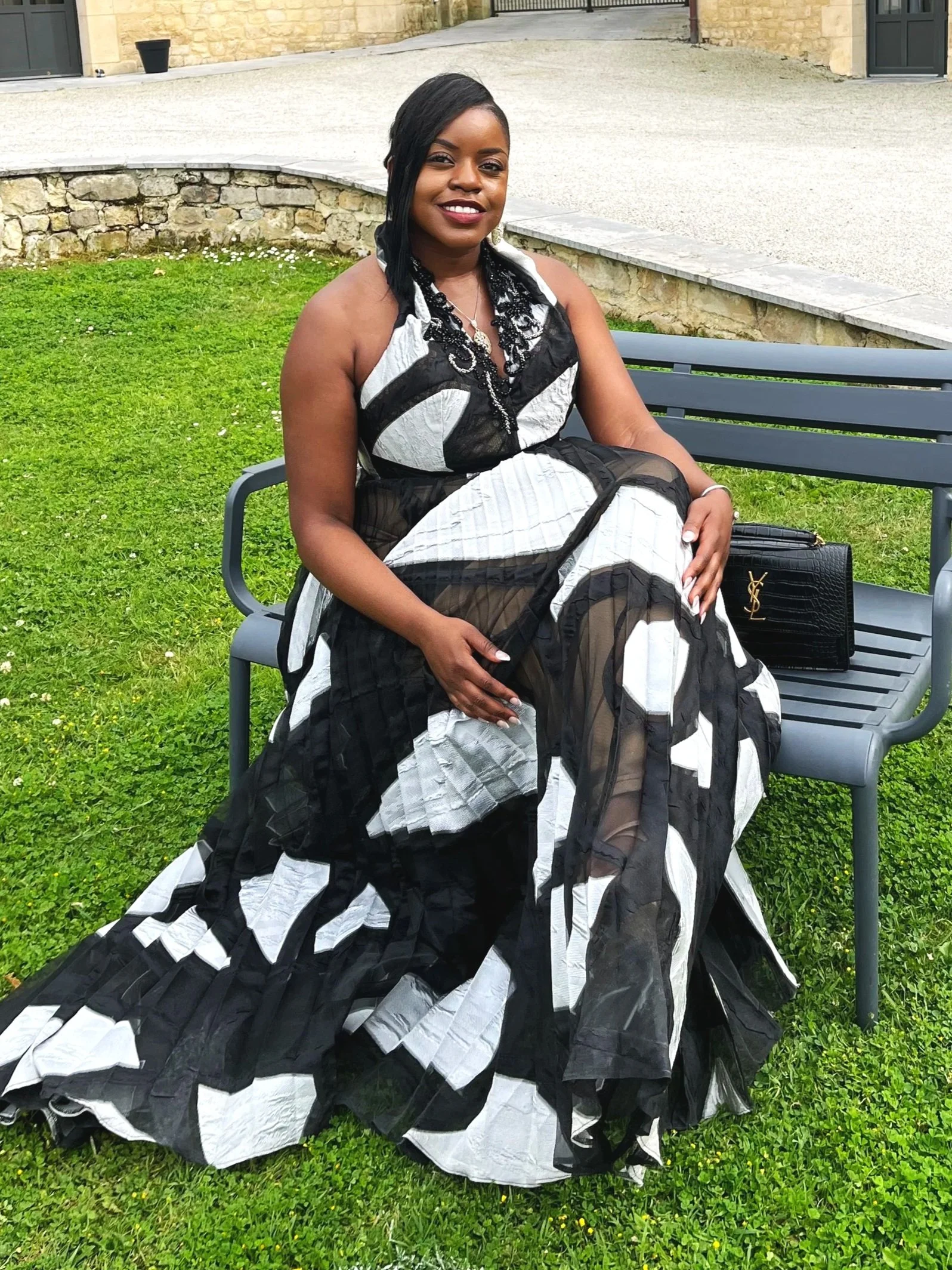 Femme assise sur un banc en extérieur, portant une robe noire et blanche à motifs. Elle a de longs cheveux noirs et sourit, avec un sac à main noir à ses côtés.