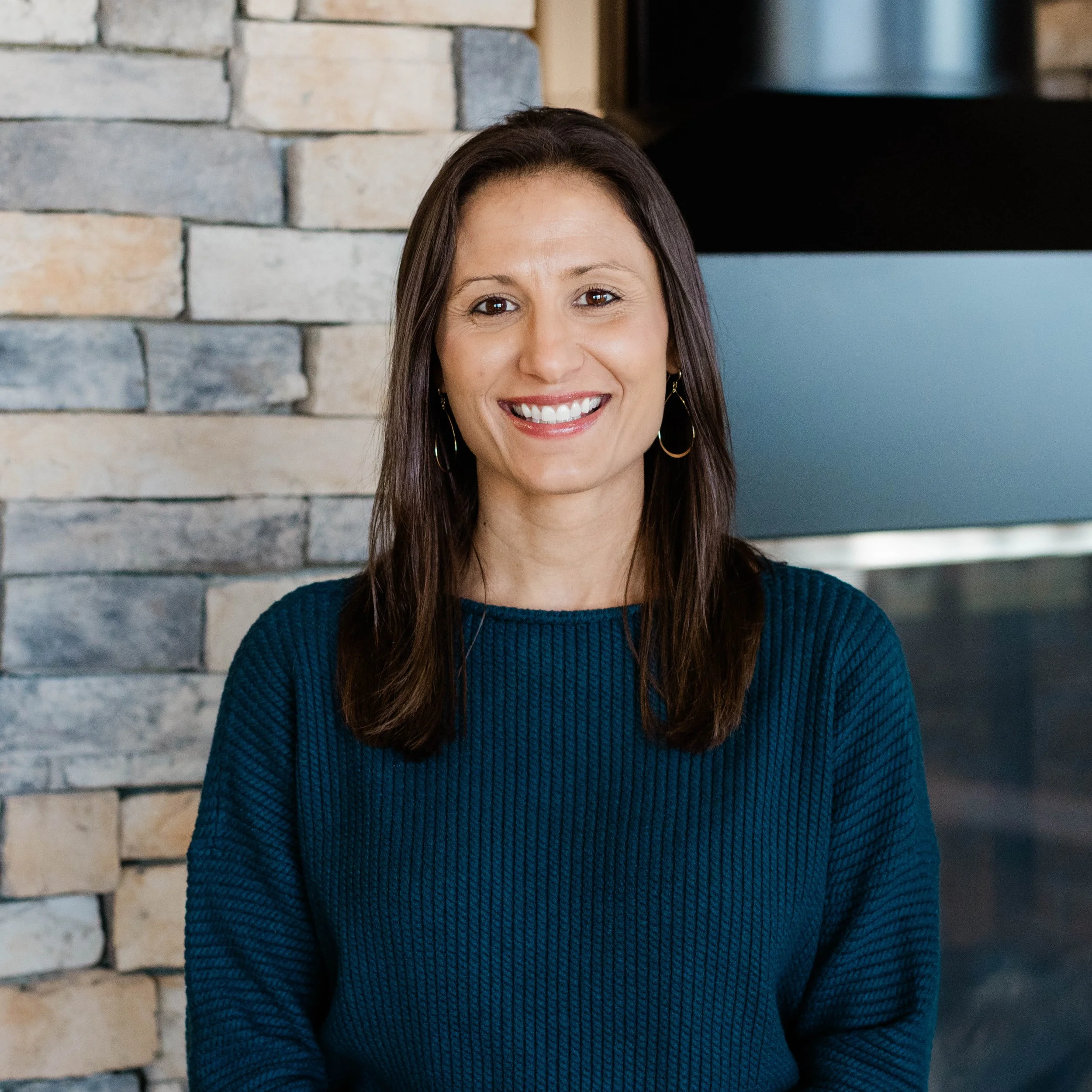Woman with shoulder-length brown hair smiling, wearing a black top, standing near a stone fireplace.