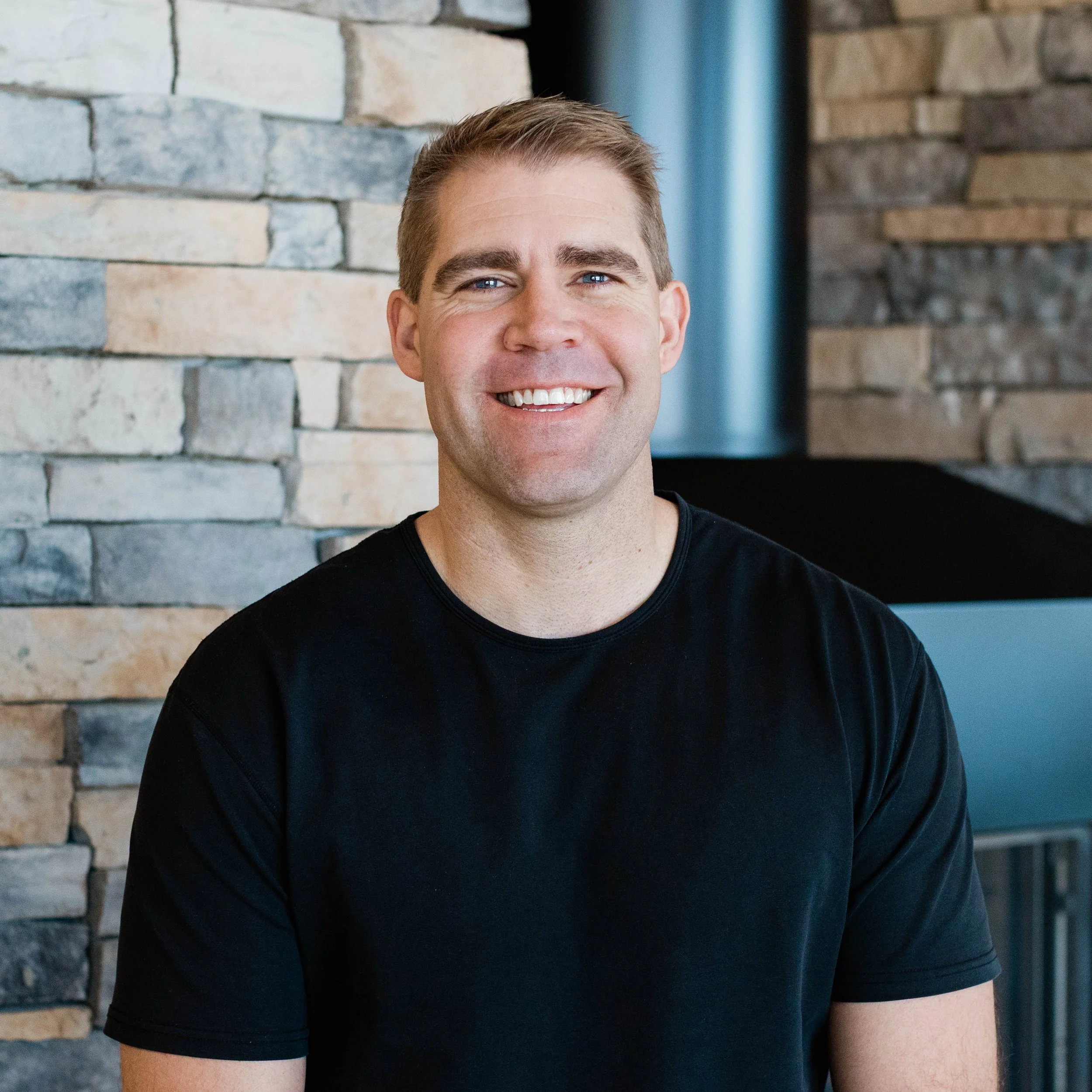 A smiling man with short, light brown hair wearing a black t-shirt, standing in front of a textured stone wall in a cozy indoor setting.