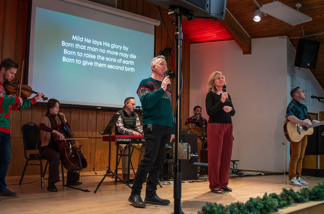 A church or community hall decorated for Christmas with a wooden interior and a stage. Four musicians are playing instruments, including a violin, cello, keyboard, and guitar. Two people are singing into microphones in front of a large screen displaying the lyrics of a Christmas song that reads: "Mild He lays His glory by, Born that man no more may die, Born to raise the sons of earth, Born to give them second birth." The stage is decorated with green garland at the front.