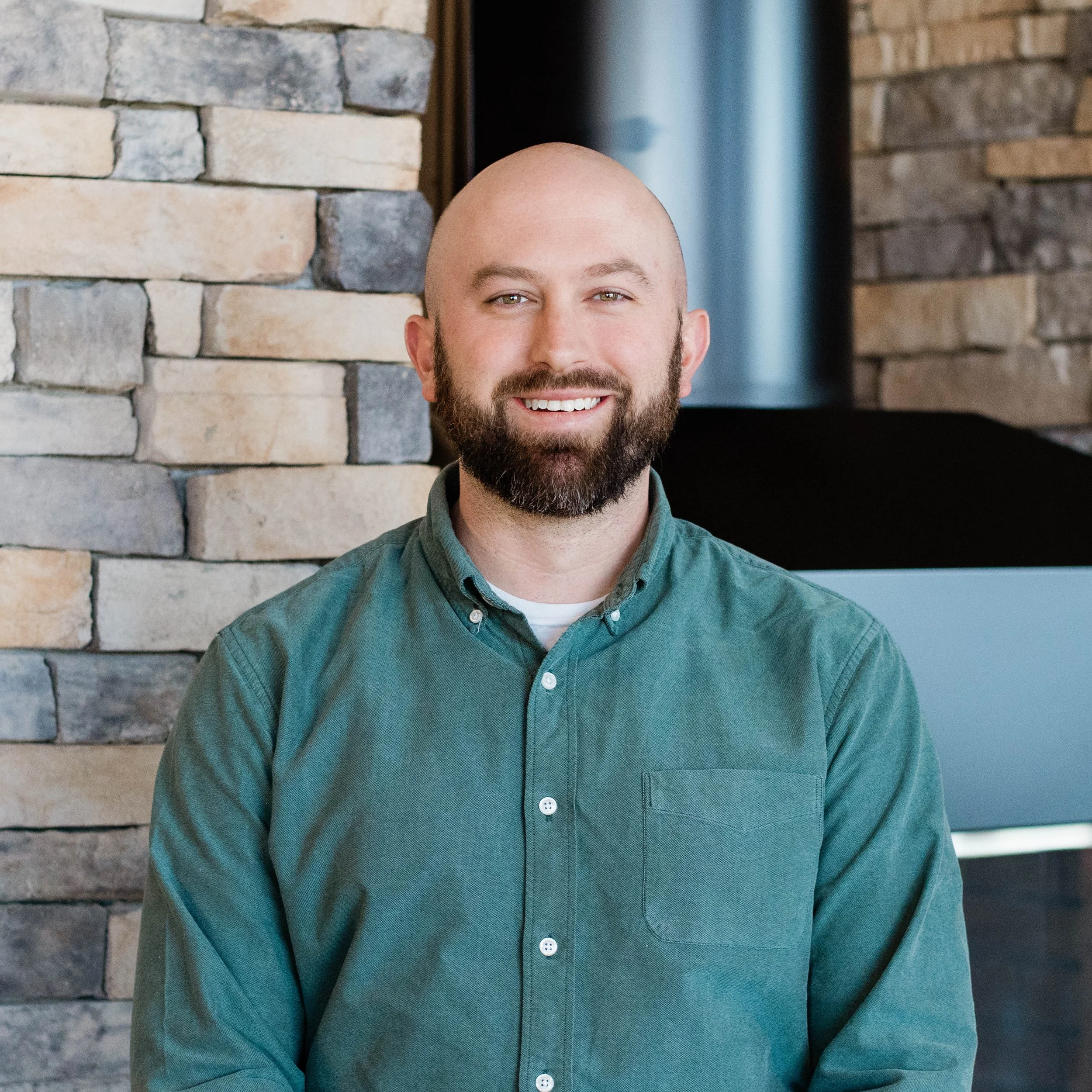 A smiling man with a beard and bald head, wearing a green button-up shirt, standing in front of a stone wall.