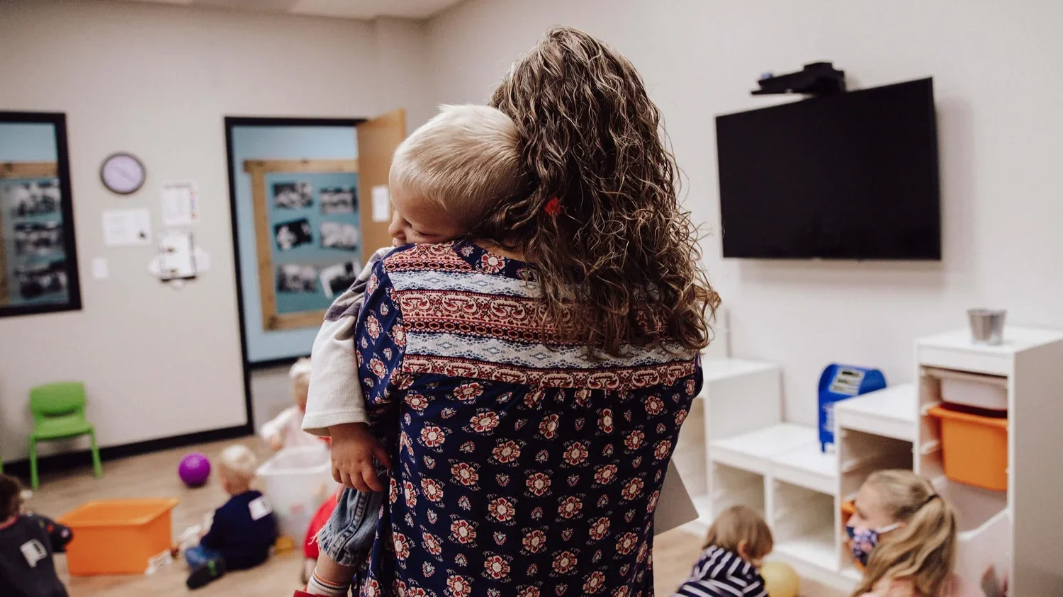 A woman with curly hair holding a young boy, hugging him in a classroom with children playing on the floor.
