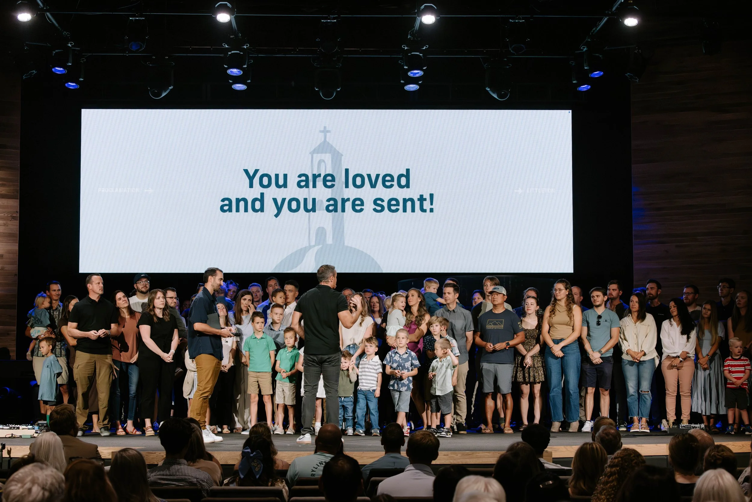 A large group of people, including children and adults, standing on stage in front of a large screen that displays the message "You are loved and you are sent!" The auditorium is filled with seated attendees watching the presentation.